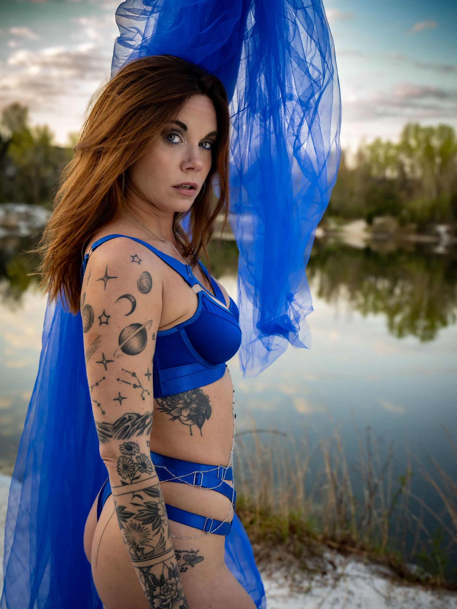 A model in a flowing blue dress poses artistically by a lake at sunset, showing off intricate arm tattoos.