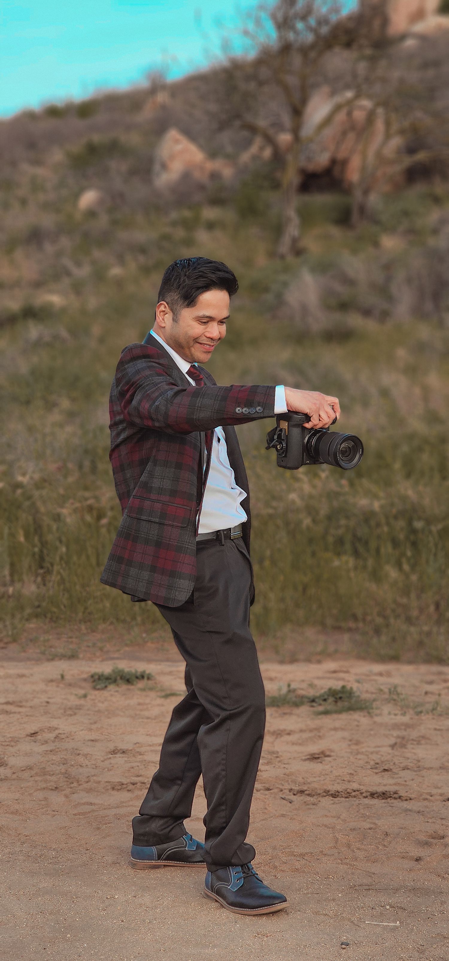 A smiling man in a plaid blazer holds a DSLR camera outdoors in a desert landscape with rocky hills behind him.