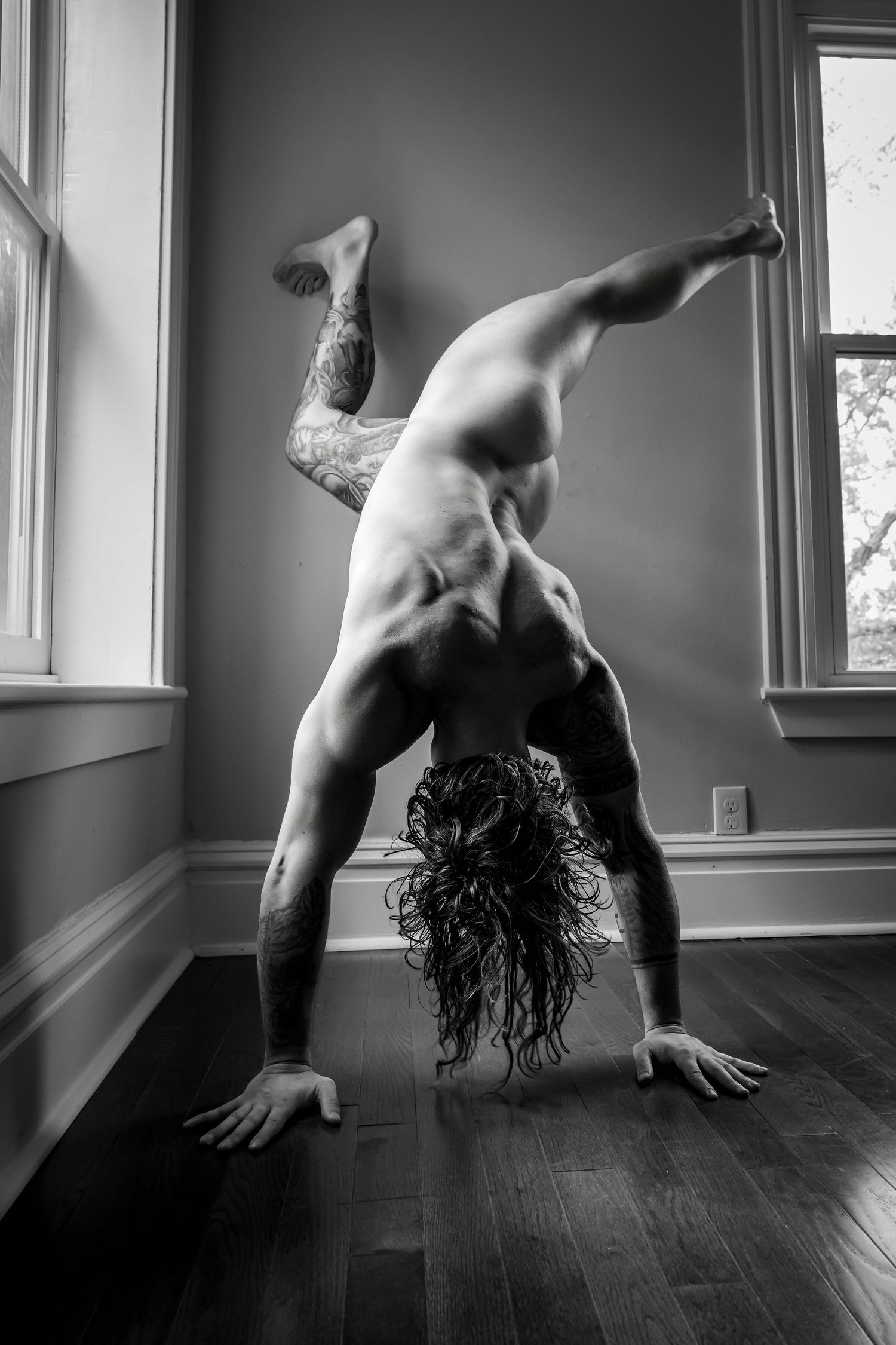 Black and white photo of a tattooed man performing a one-handed handstand on hardwood floors near windows.