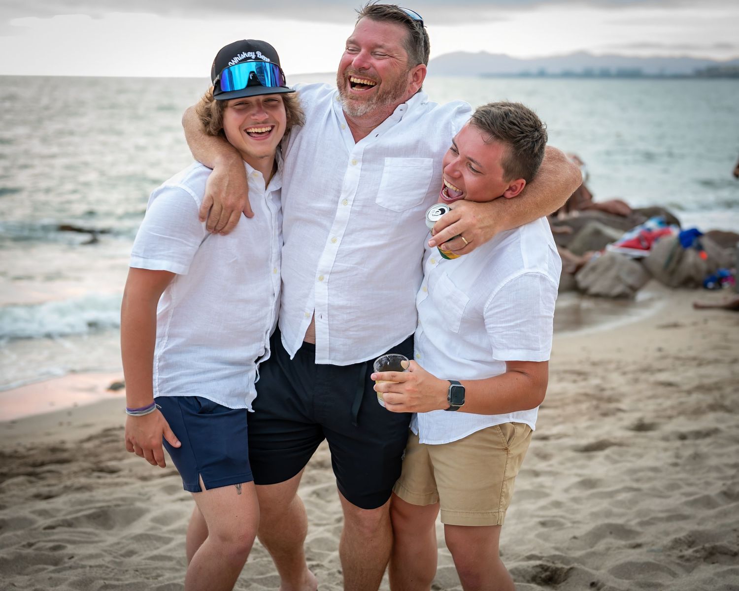 Three people in white shirts sharing joyful embrace on beach at sunset.