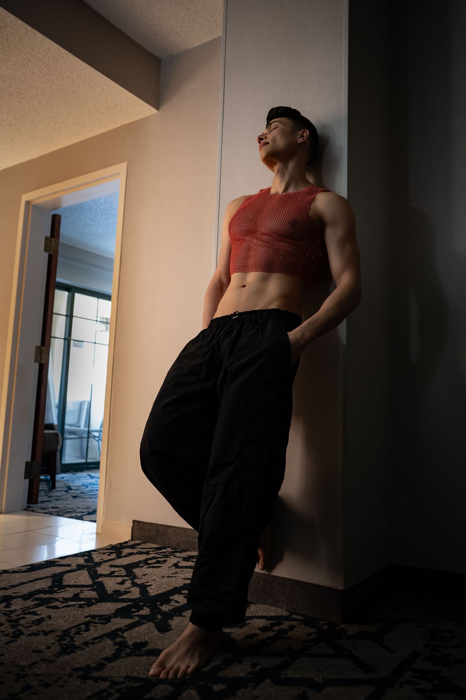 Fit young man in red crop top and black pants leans against wall indoors, looking up confidently.