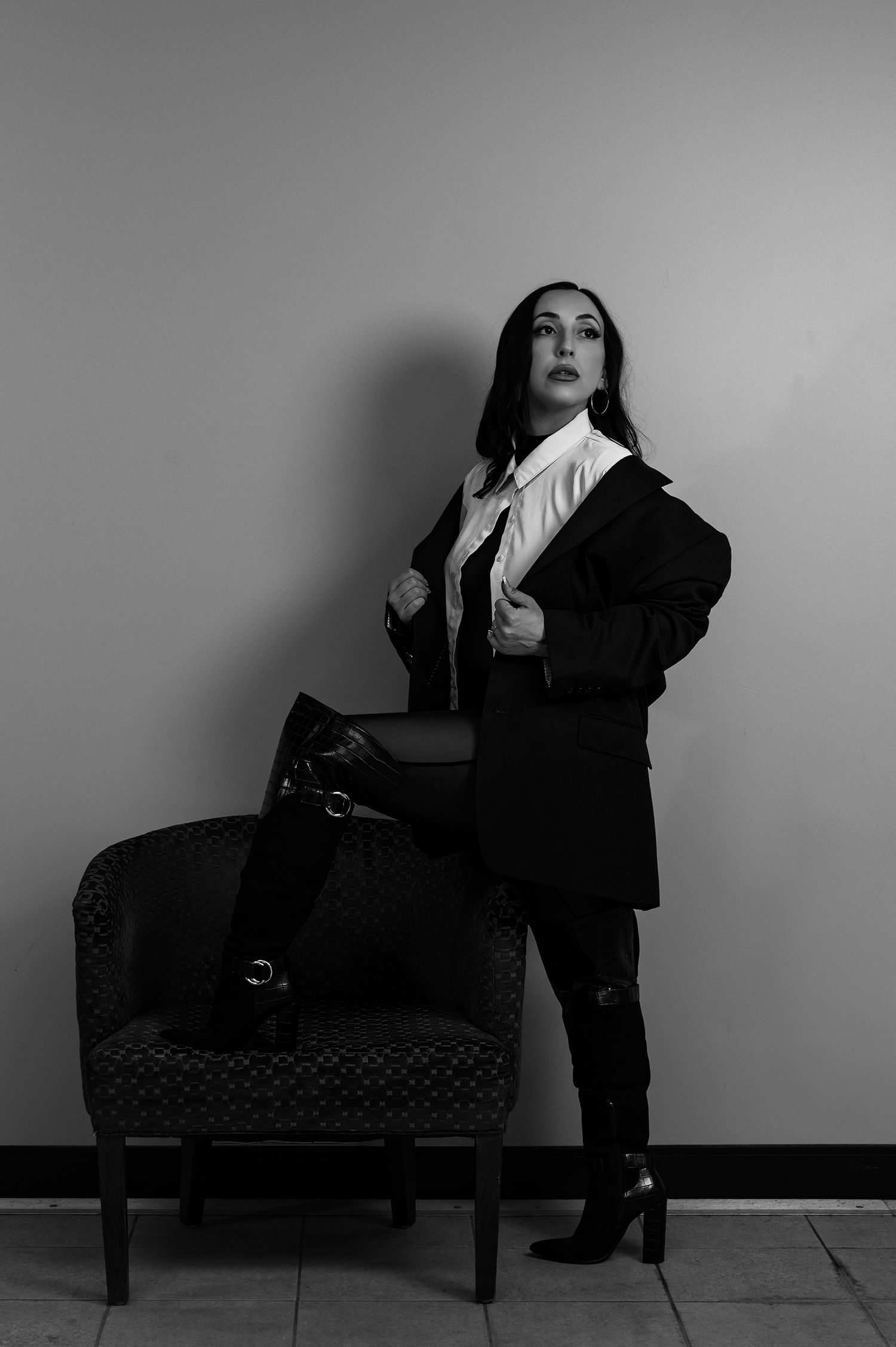 Black and white portrait of a woman in a blazer and boots posing confidently beside an armchair.