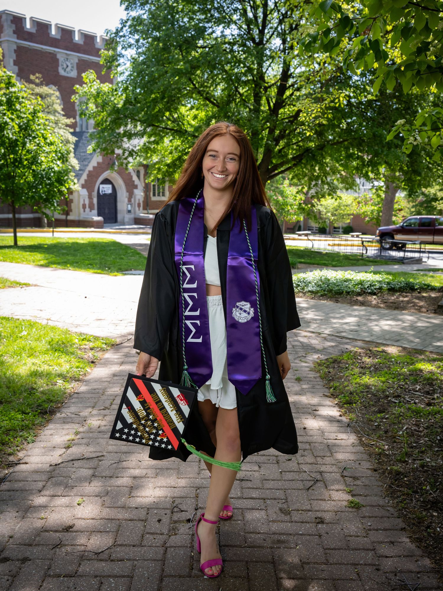 Graduate wearing black cap and gown with purple sorority stole poses on brick pathway surrounded by trees.
