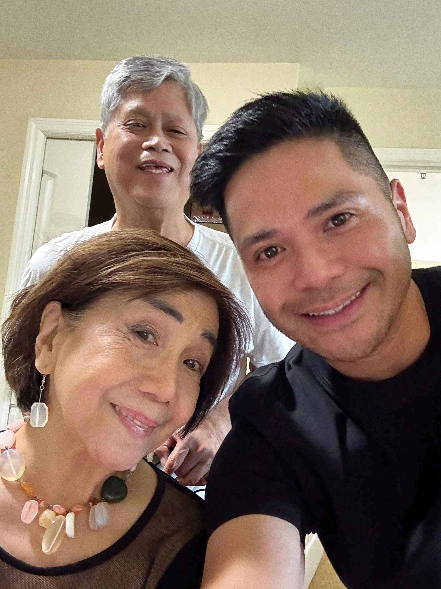 Three people smiling together for a selfie, two older adults and one younger man in a black shirt.