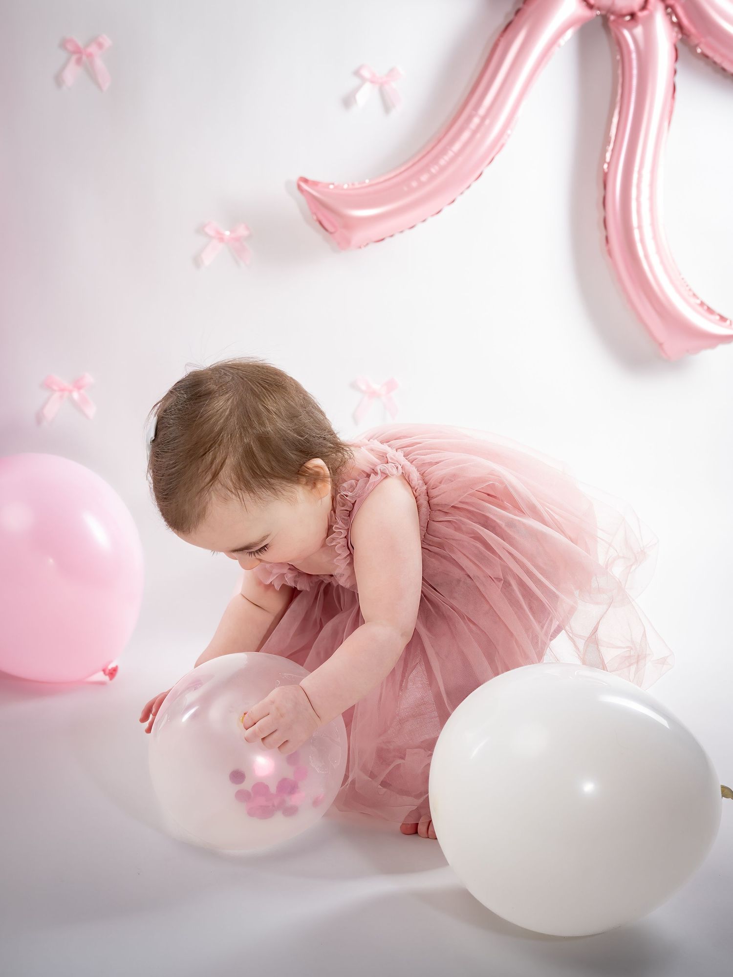 Baby in pink dress playing with balloons during first birthday photoshoot.