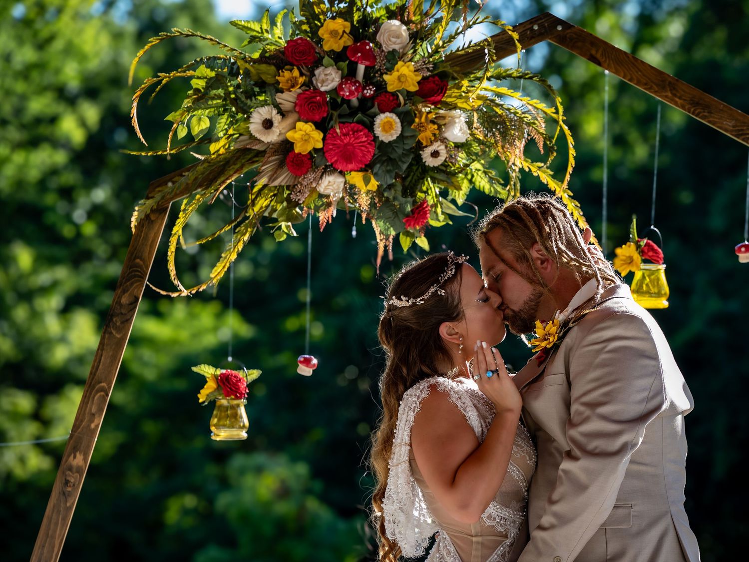 Romantic wedding kiss under a wooden arch adorned with colorful wildflowers and hanging lanterns.
