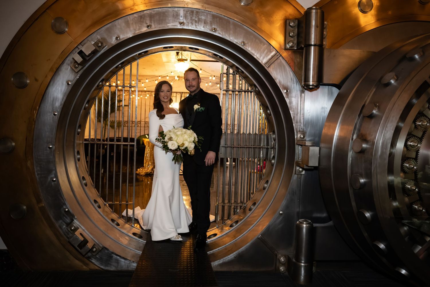 Wedding couple photographed through vintage bank vault door frame with dramatic lighting.