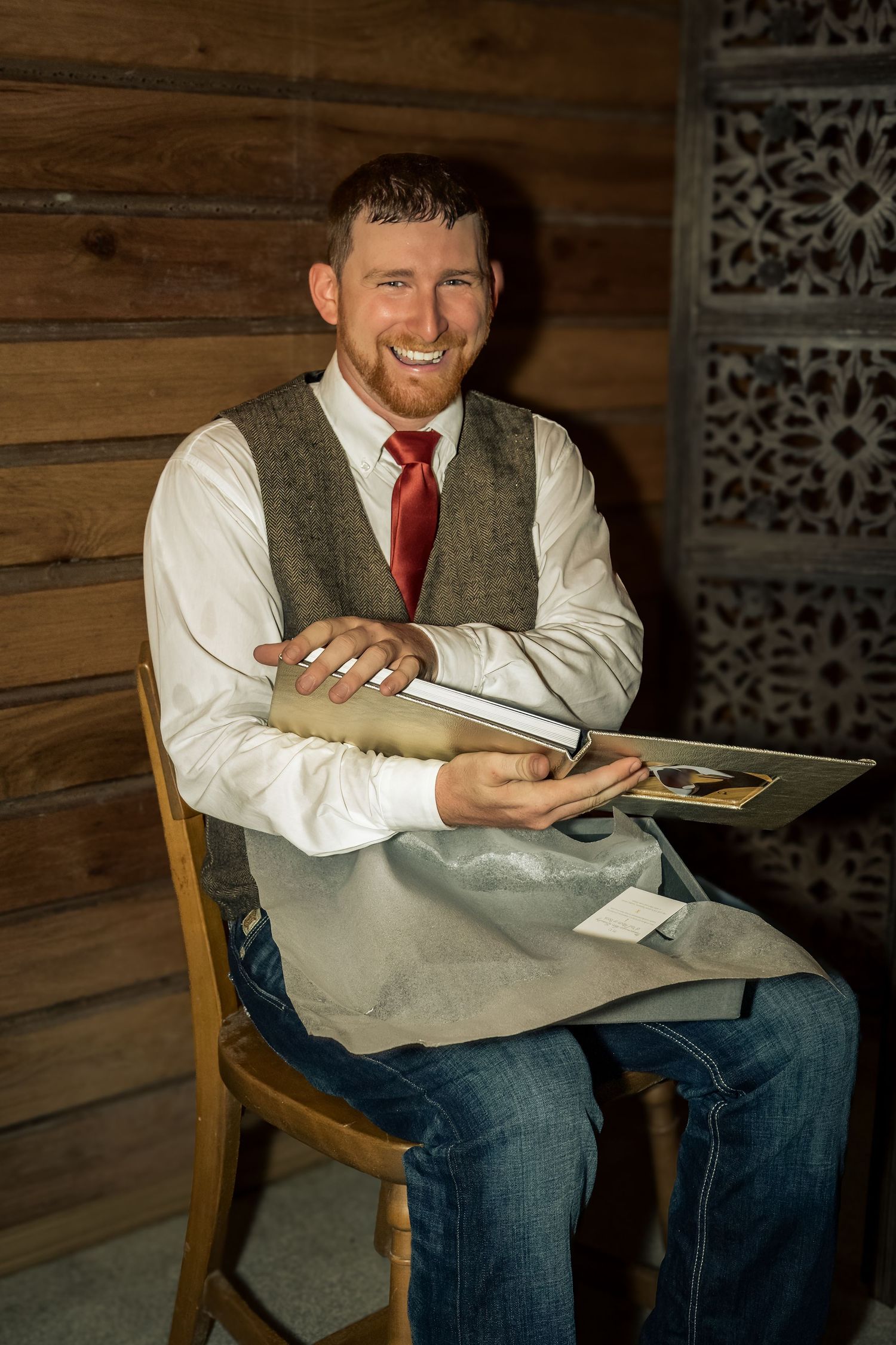 Smiling man in vest and red tie sits on chair holding a photo album in a rustic wood-paneled room.