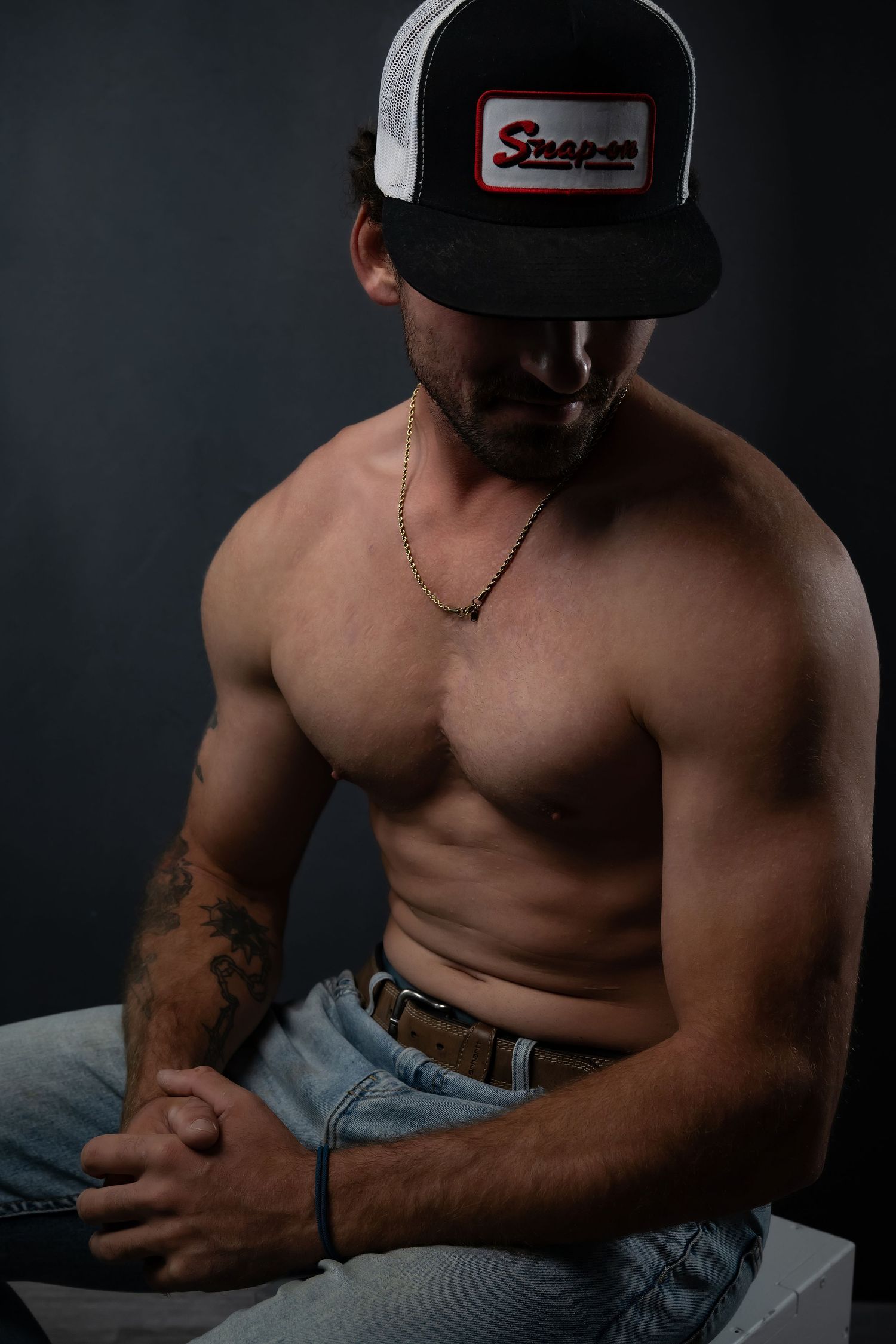 Muscular shirtless man wearing a Snap-on trucker hat and jeans, sitting with tattoos visible, against a dark background.
