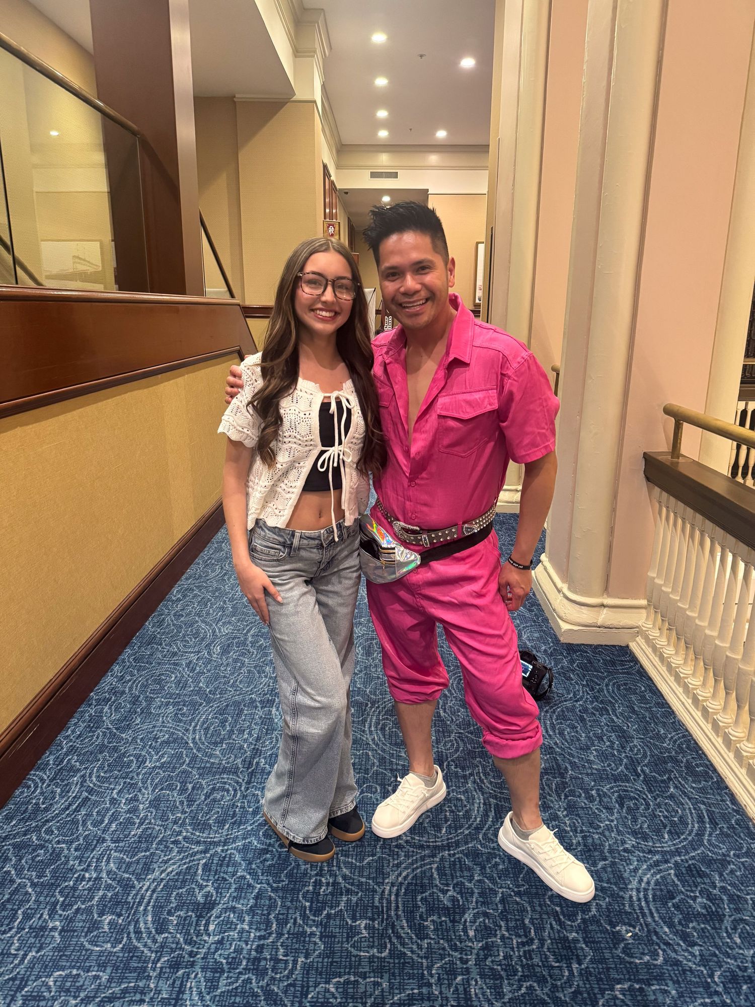 Two people posing together in a hotel hallway, one wearing a bright pink outfit and the other in jeans and white top.