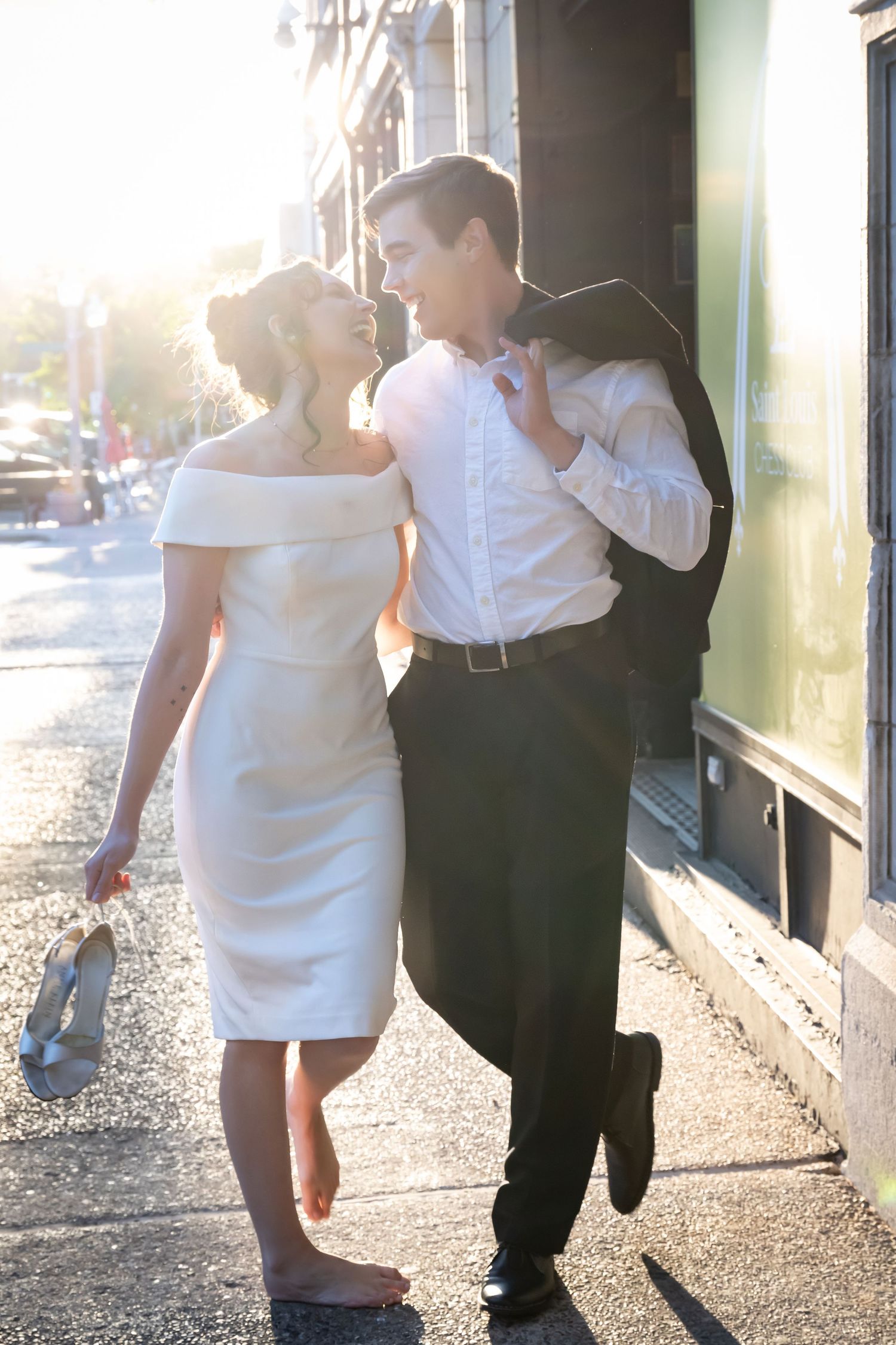 Couple in wedding attire sharing a romantic moment on a sunny city sidewalk, bride in white dress holding shoes.