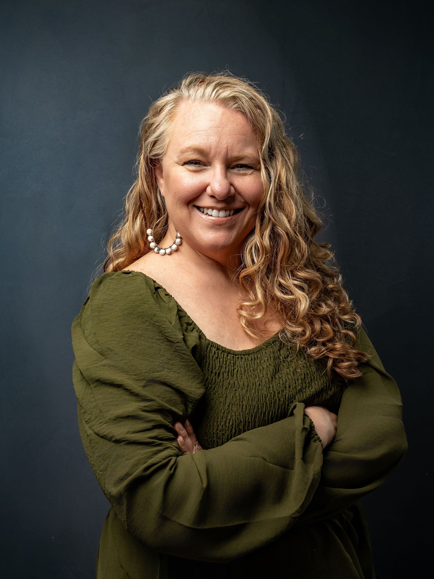 Smiling woman with curly blonde hair in green top, arms crossed, professional studio portrait photo.
