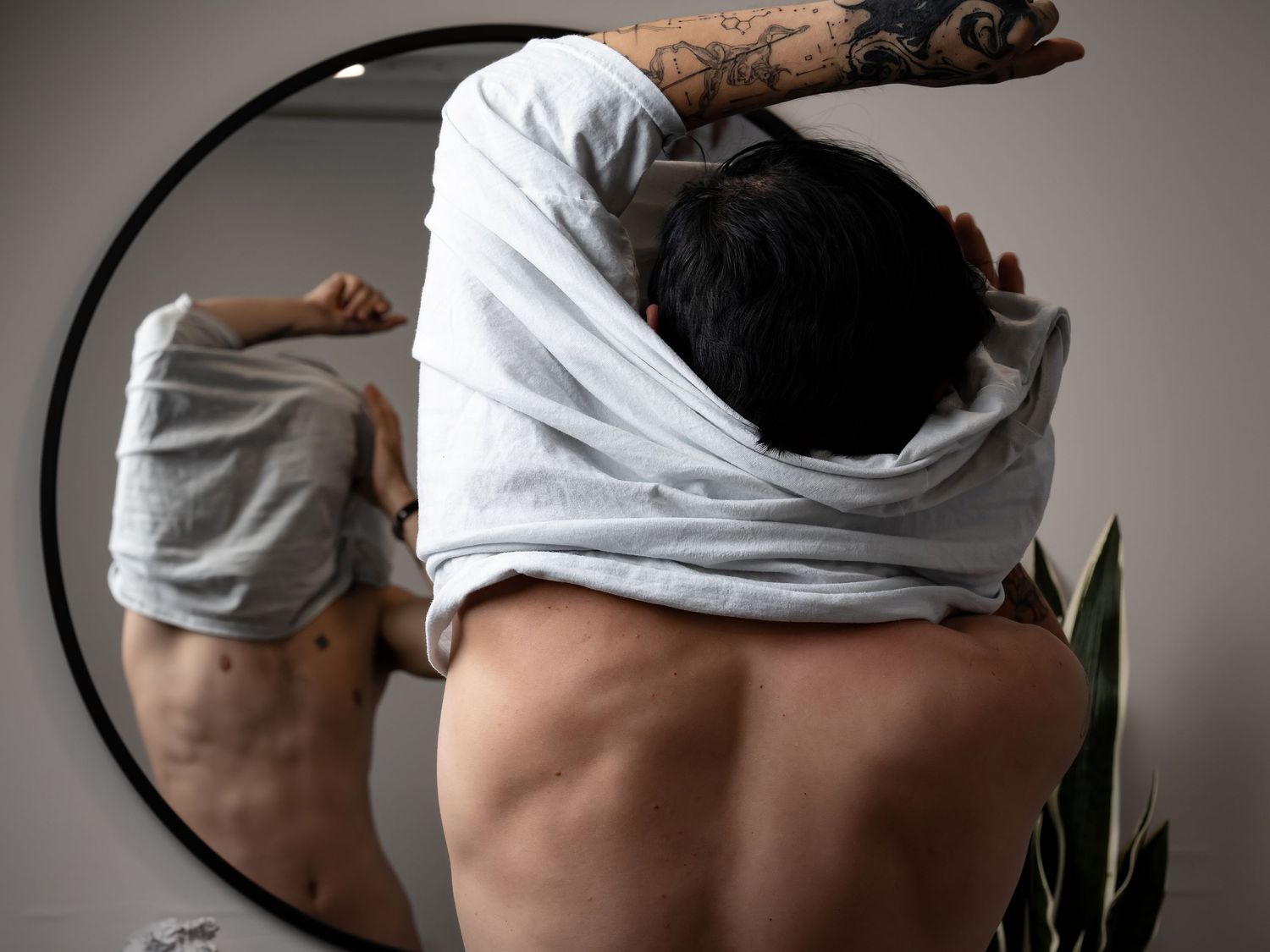 Man removing gray shirt in front of round mirror, revealing tattooed arm and muscular back, with plant in background.