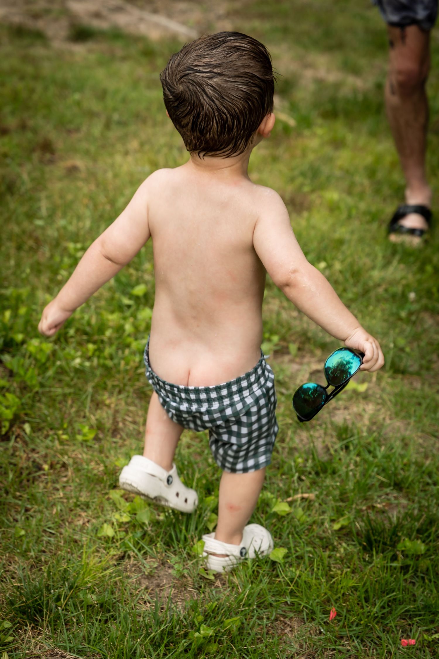 Toddler in checkered swim shorts running through grass holding blue toy.
