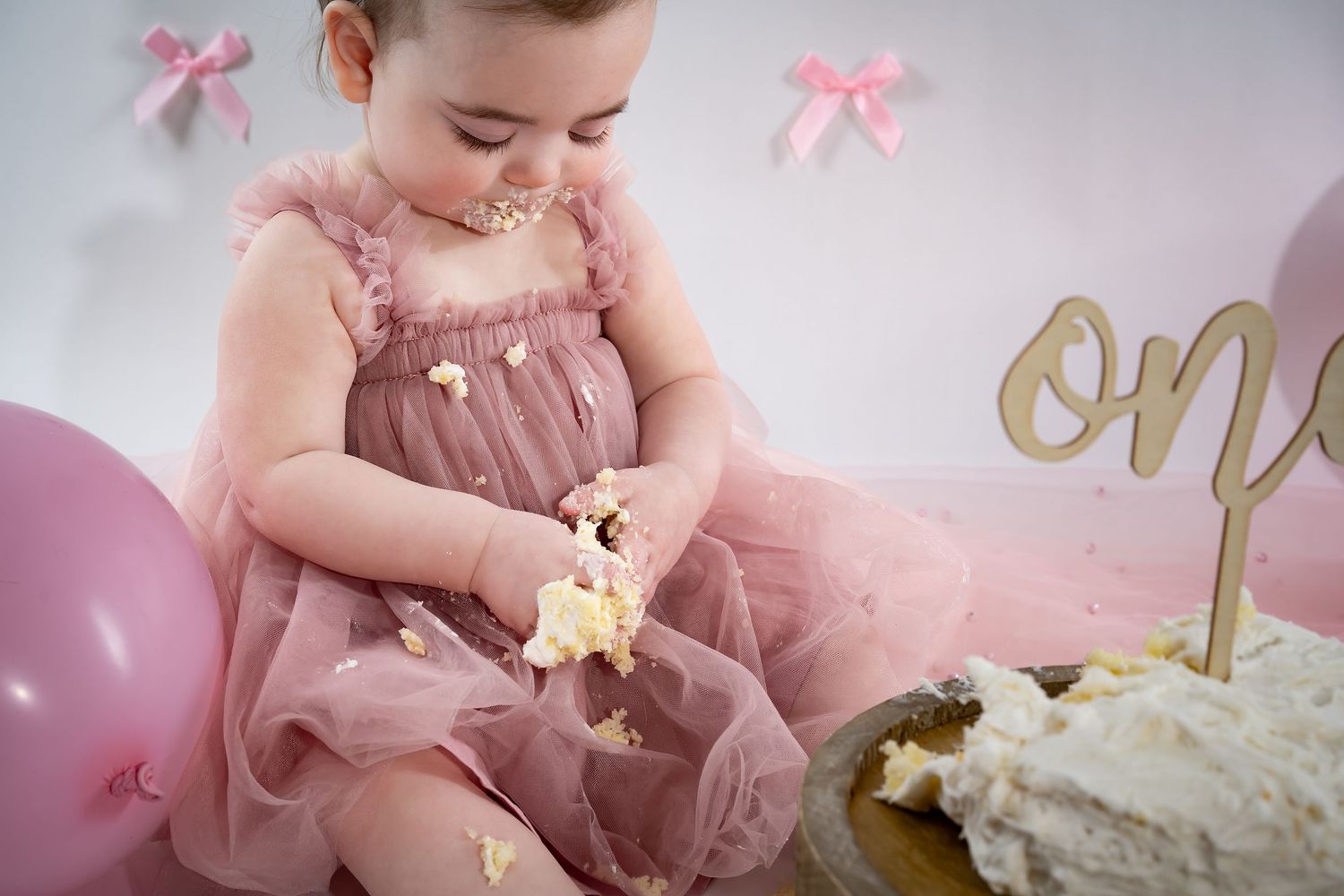 Baby in pink tulle dress reaching for birthday cake with gold decorations and pink balloons.
