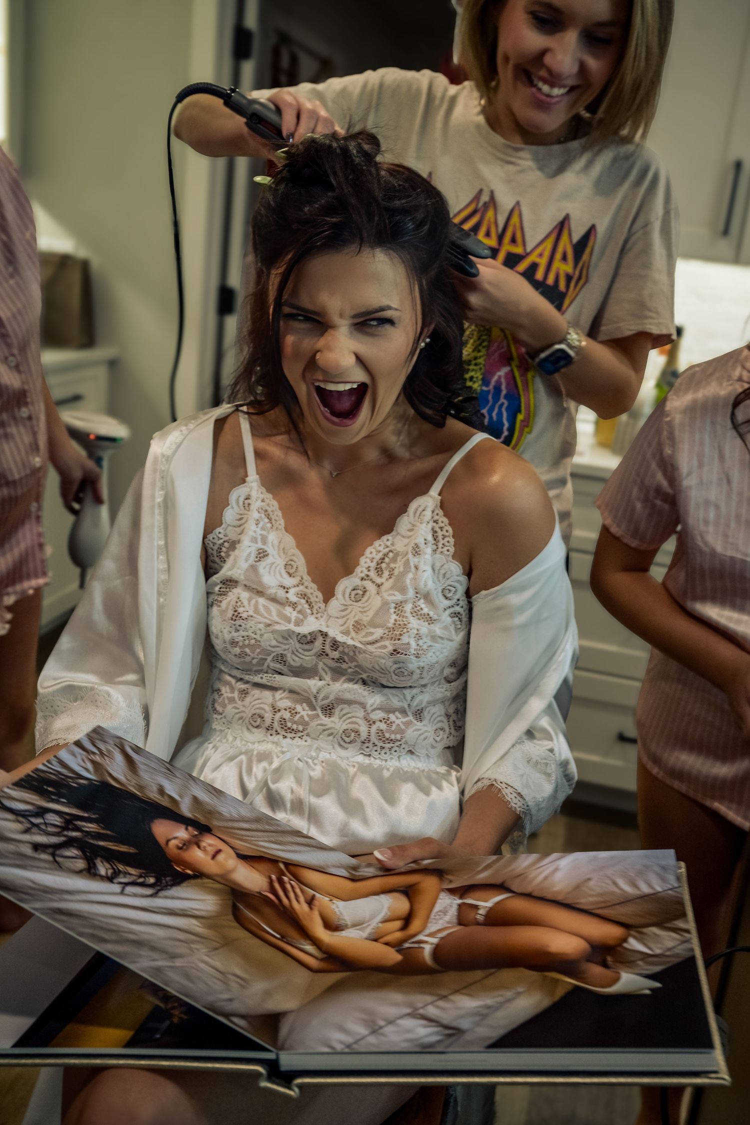 Bride in white lace dress laughs while hairstylist uses curling iron during wedding preparations.