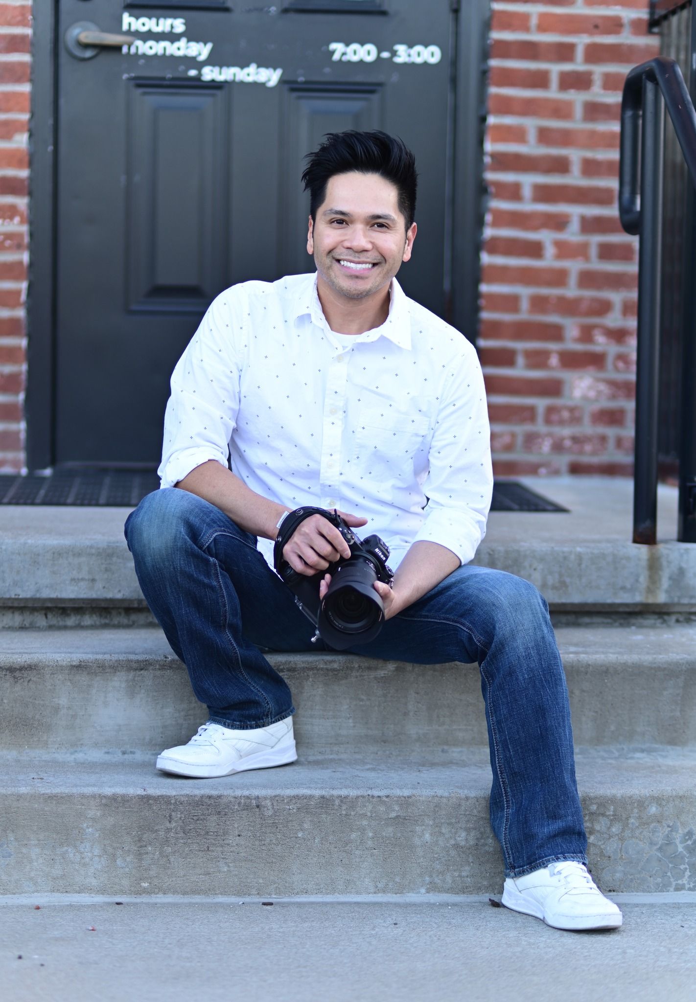 A photographer in a white button-down shirt and jeans sits casually on concrete steps holding a camera.