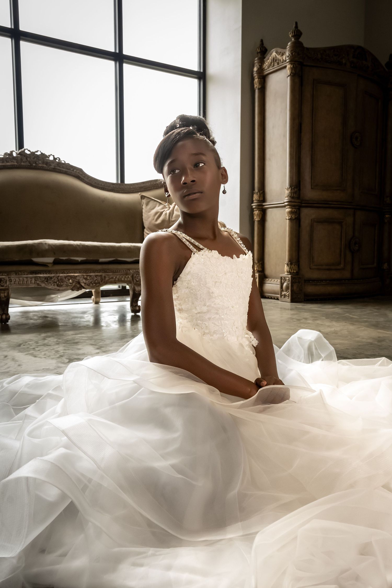 A bride in a flowing white gown reclines elegantly on the floor near large windows.