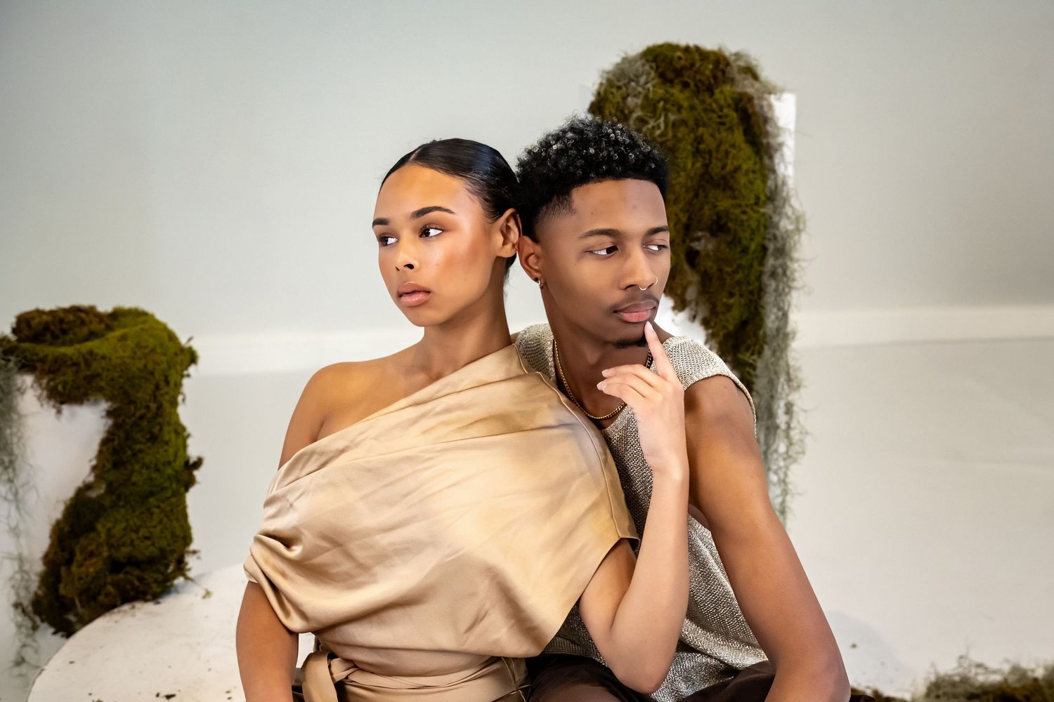 Two models pose artistically against textured wall in beige and neutral tones.