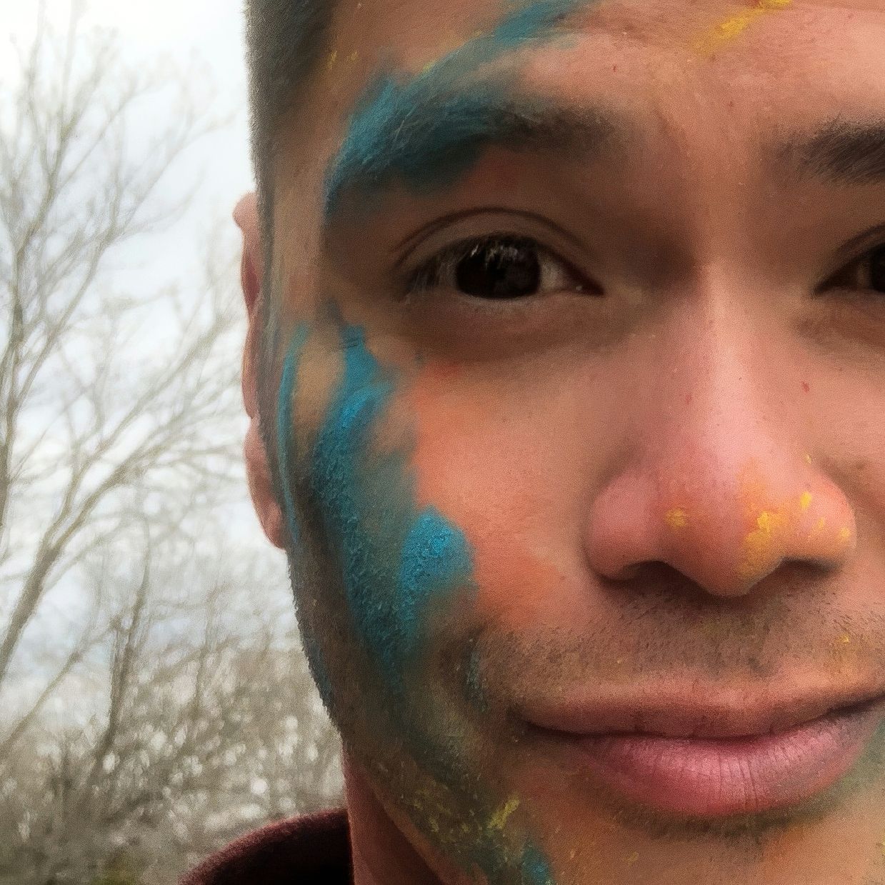 Young man with colorful blue and yellow Holi powder paint on his face outdoors with bare trees in background.