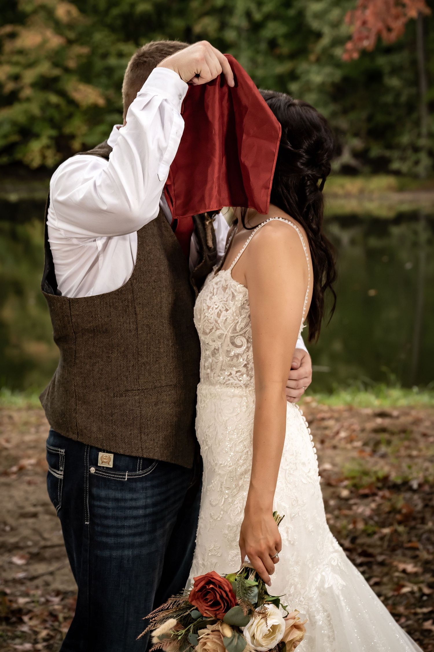 A couple in wedding attire embraces near a lake with autumn foliage in the background.