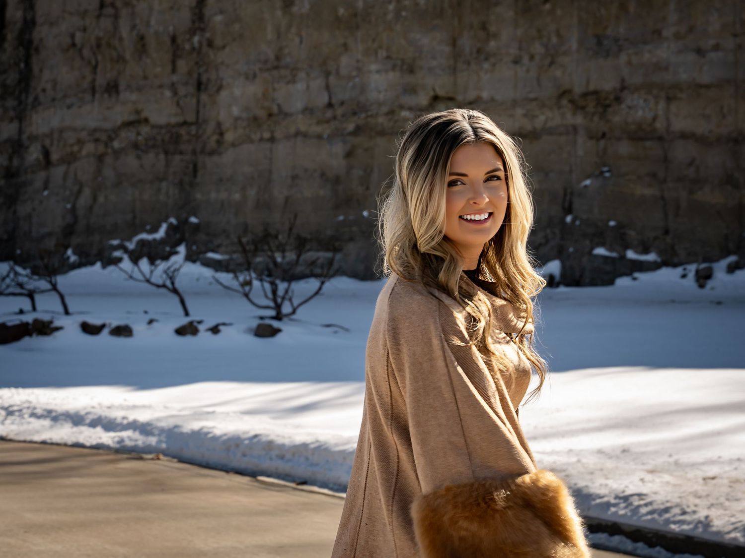 Winter fashion shoot featuring camel coat and boots against stone wall with snow.