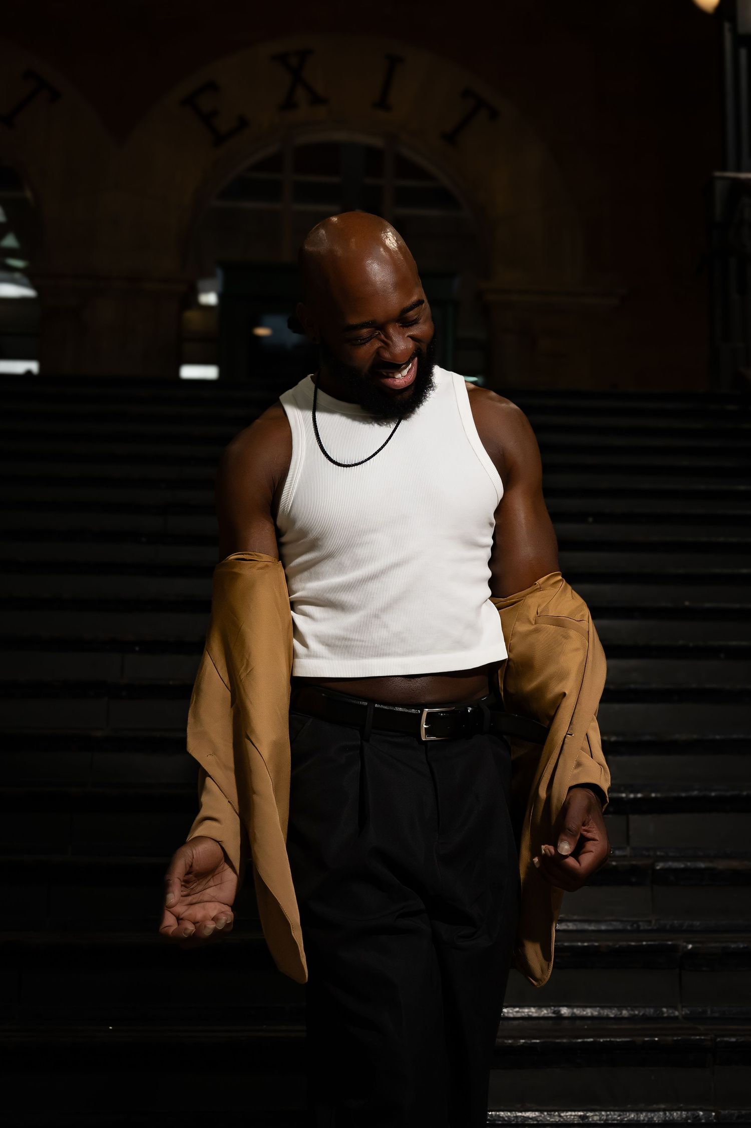 Casual urban portrait on stairs featuring white tank top and tan jacket in low light setting.