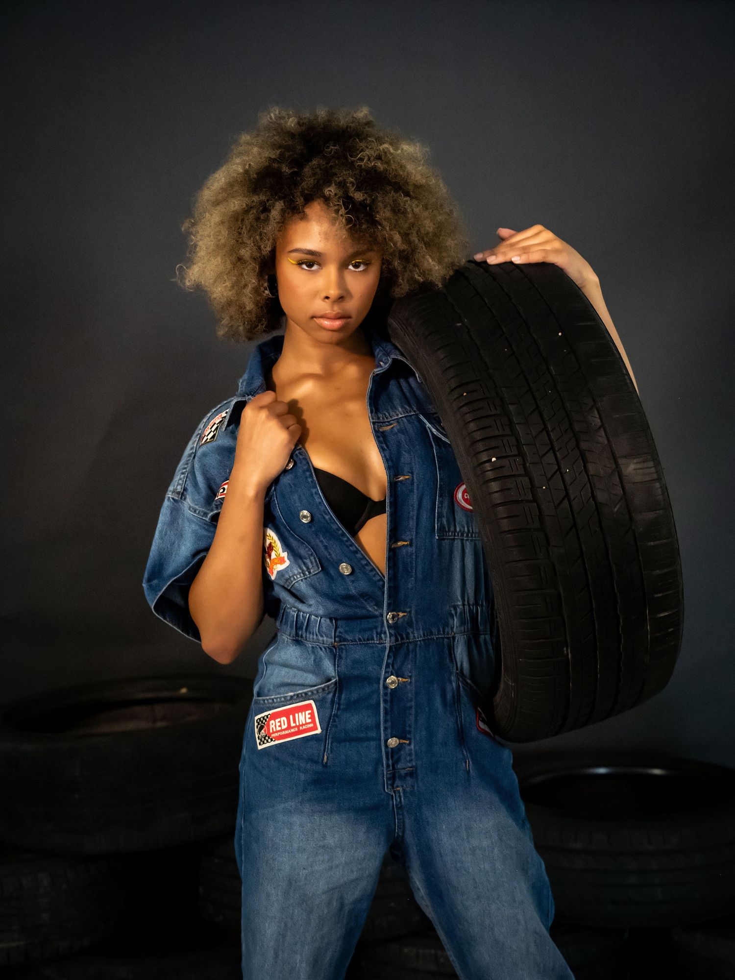 Model in denim jacket poses with car tire against dark background.