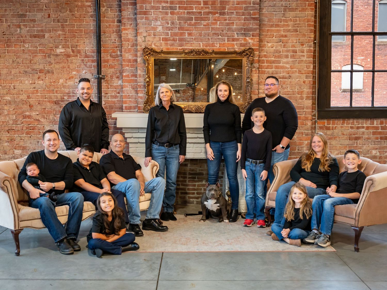 Family group portrait in casual attire seated on benches against rustic brick wall.