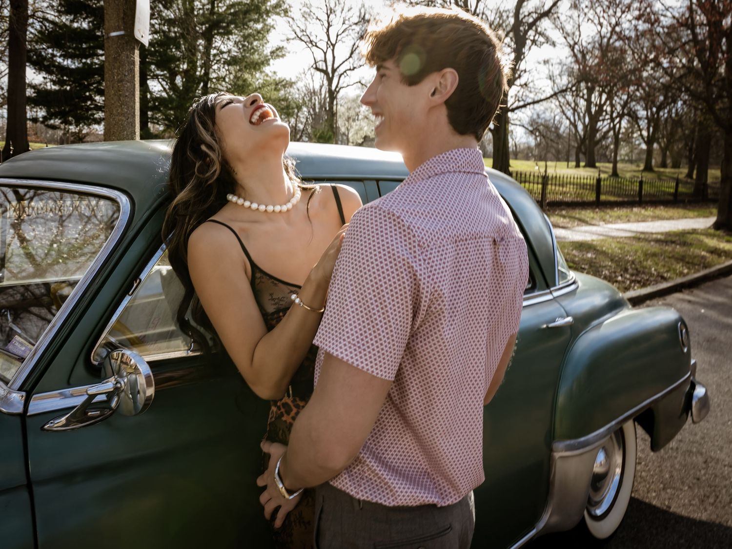 A laughing couple in vintage attire embraces beside a classic teal car in a romantic outdoor setting.