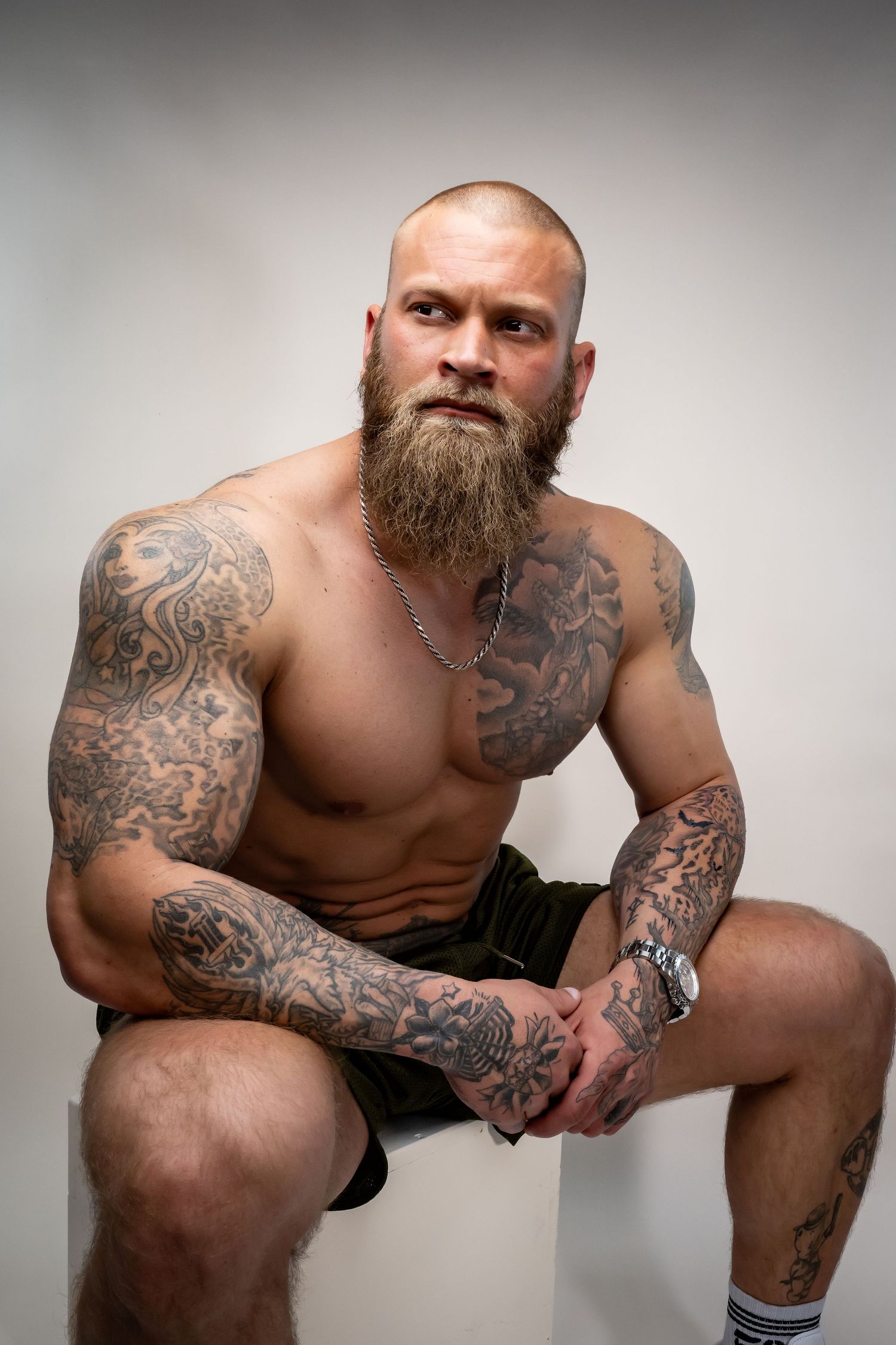Muscular bald man with full beard and extensive tattoos sits shirtless wearing dark shorts and a silver chain necklace.