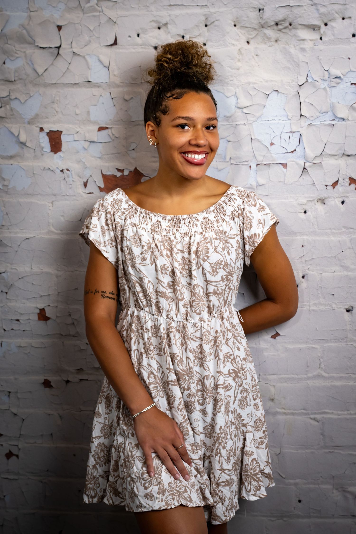 Woman in floral dress poses confidently against textured white brick wall.