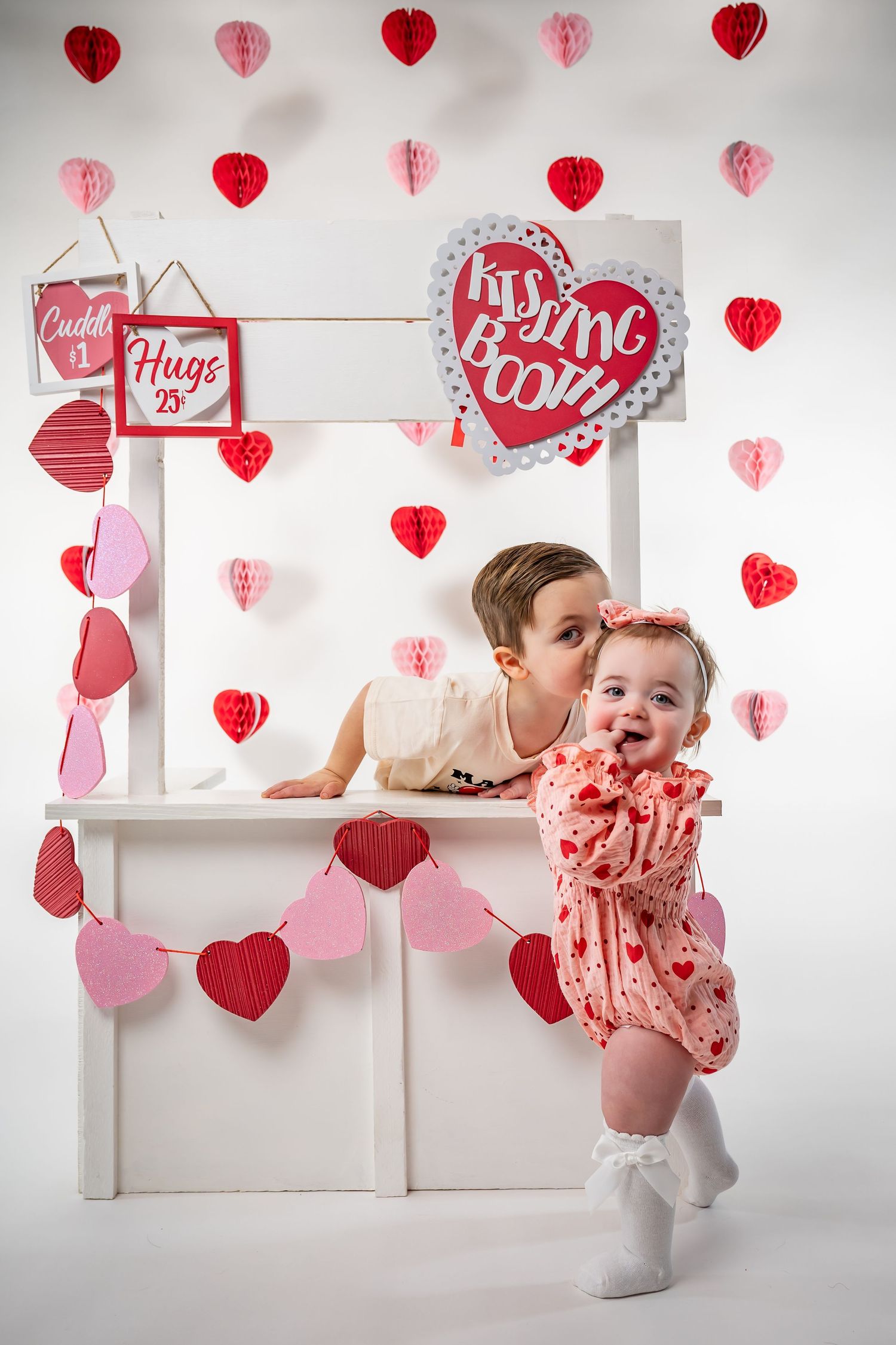 Valentine's themed photo setup with child at white kissing booth decorated with red hearts.