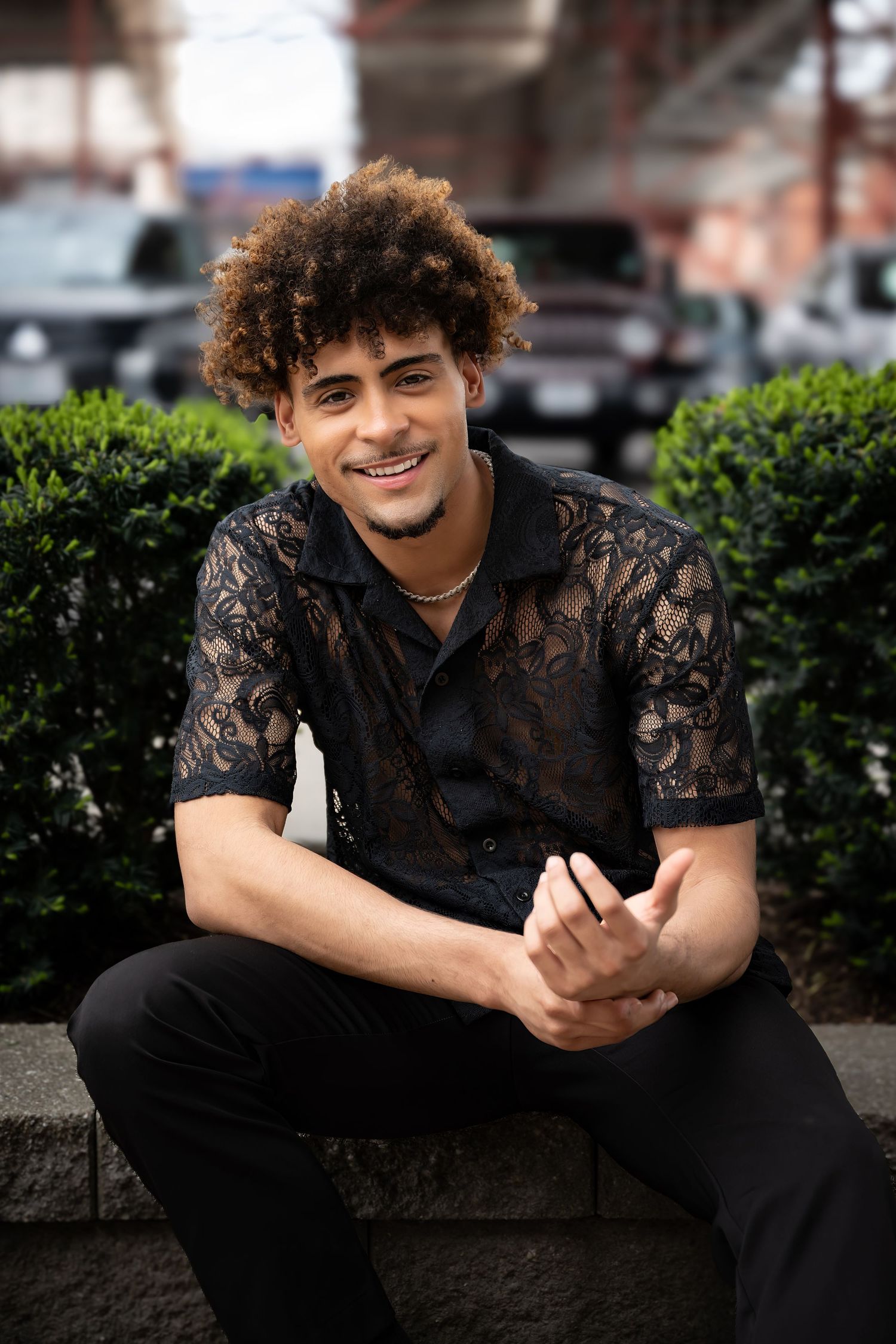 Person in black lace shirt sitting outdoors by greenery.