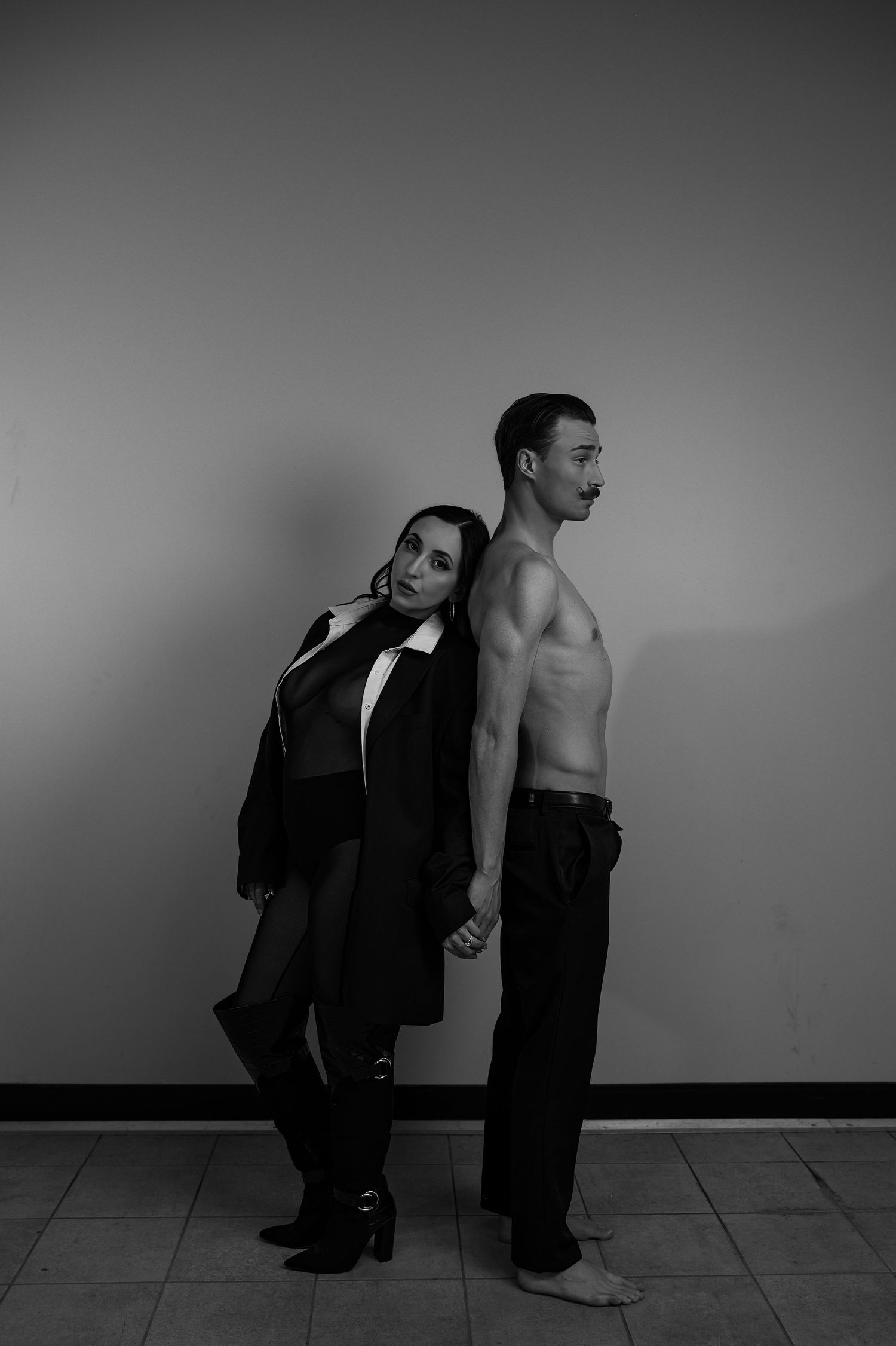 Black and white photo of a couple posing back to back, woman in blazer and man shirtless in jeans.