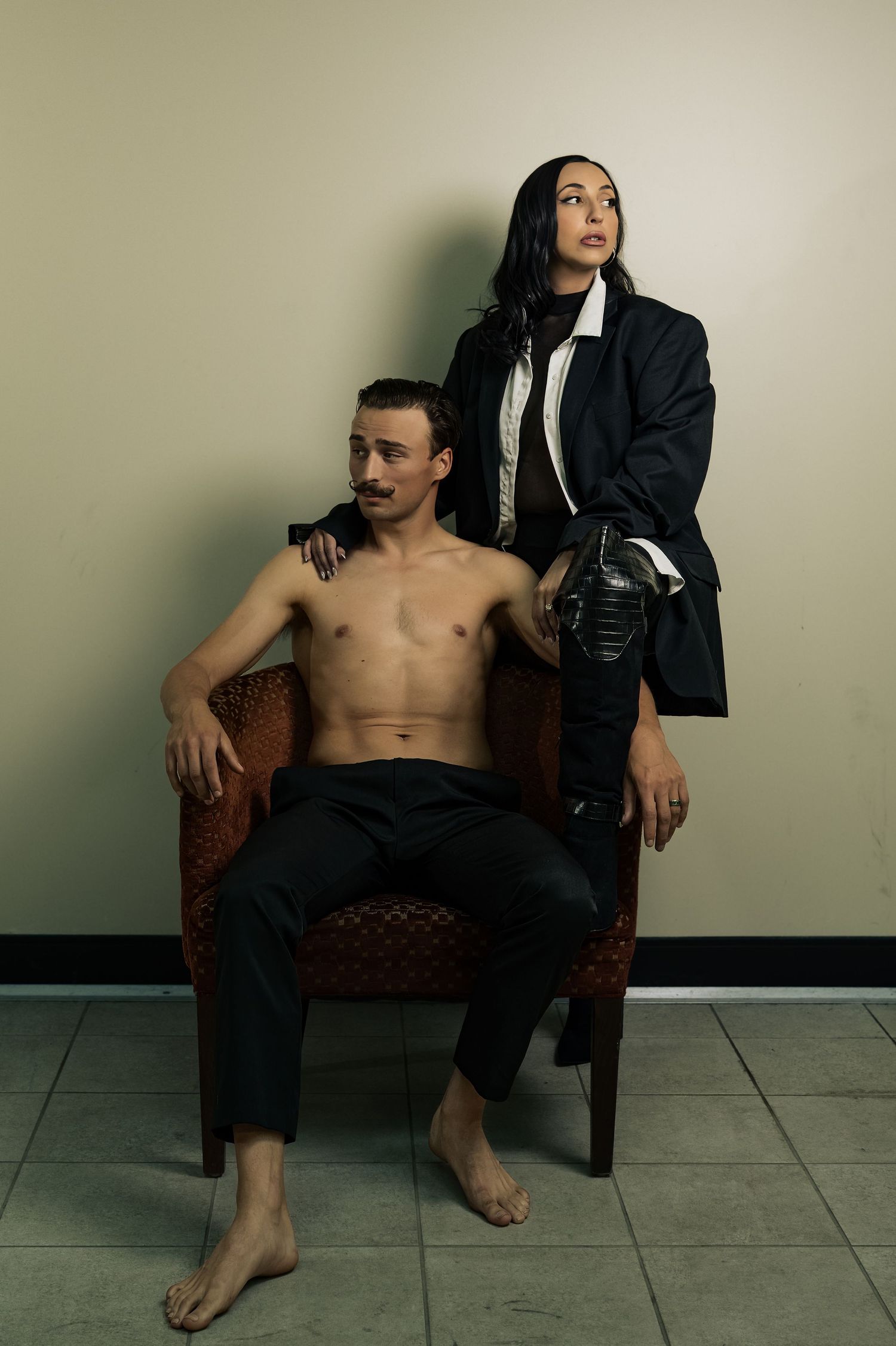 A shirtless man sits in a wicker chair while a woman in a black blazer stands behind him in a stylish editorial pose.