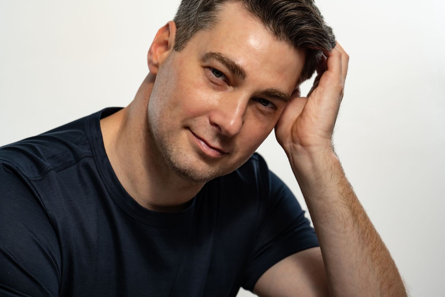 A smiling man in a dark blue t-shirt rests his head on his hand against a light gray background.