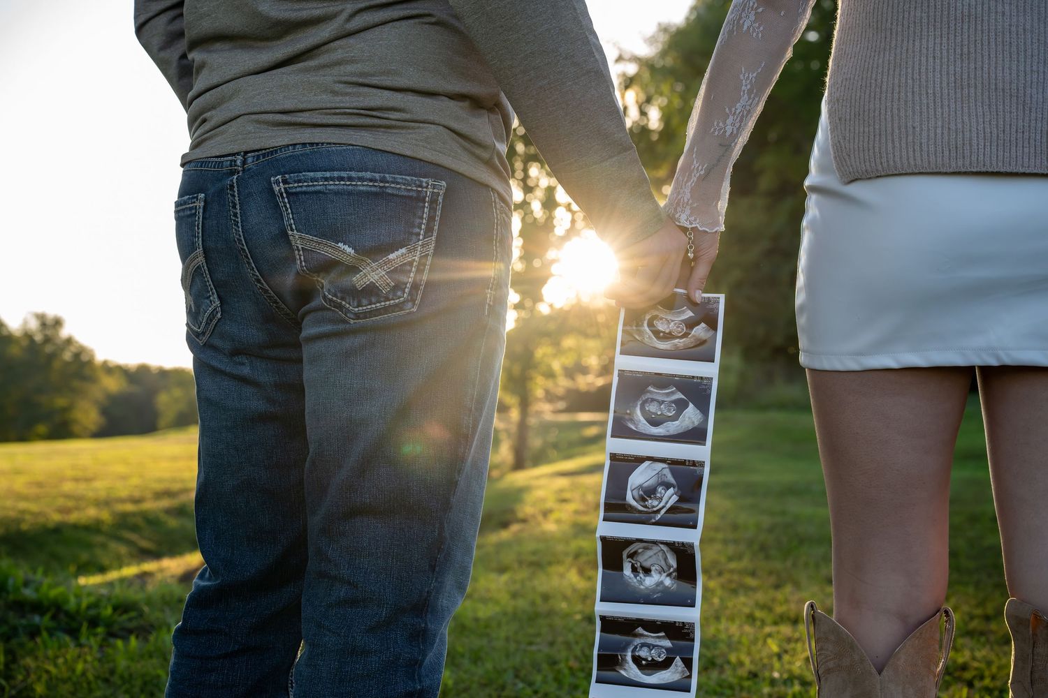 Couple holding ultrasound photos in sunlit field with pregnancy announcement strip against sunset backdrop.