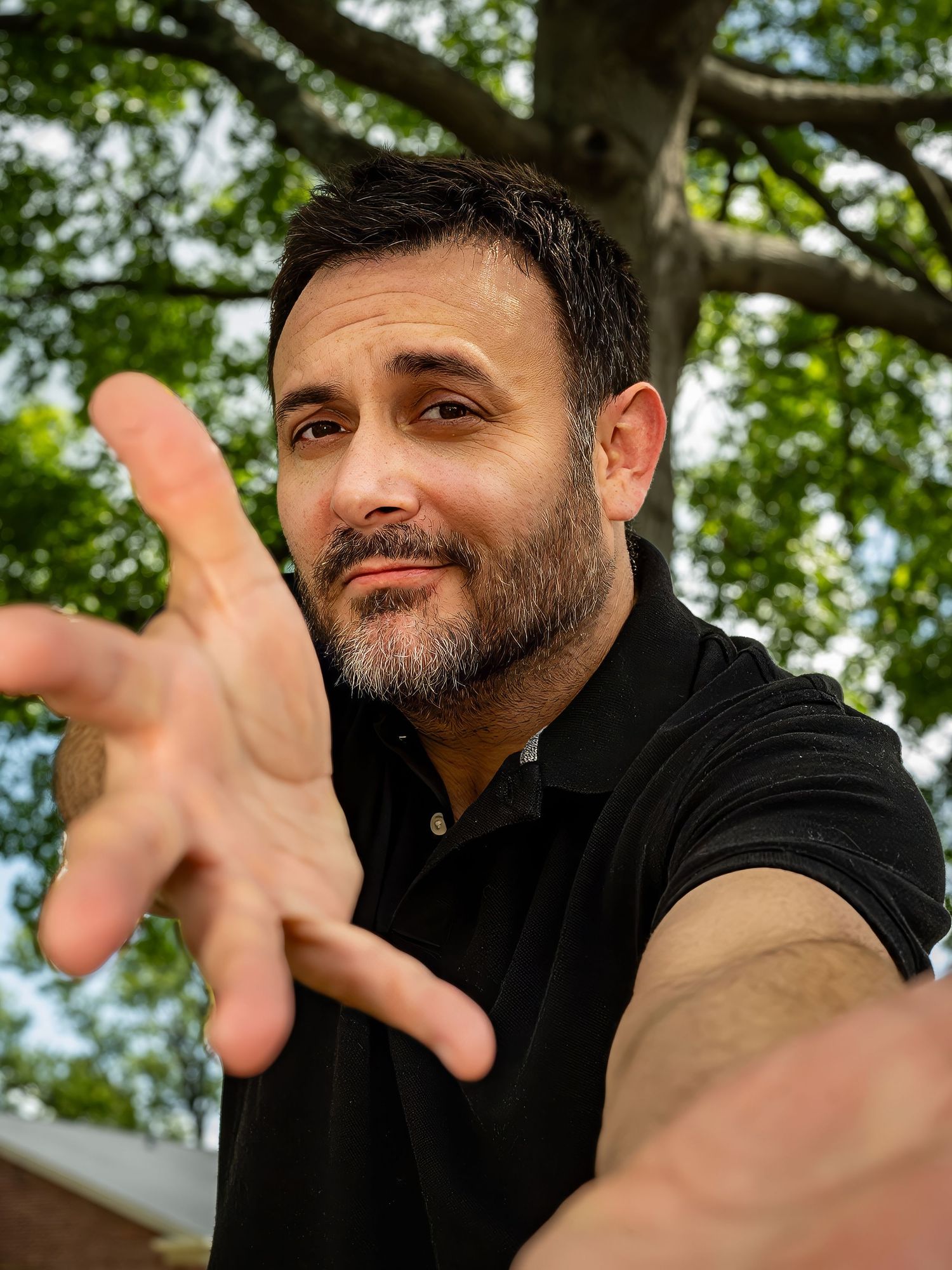 Man in black shirt reaching toward camera outdoors under green trees, with a friendly smirk and short beard.