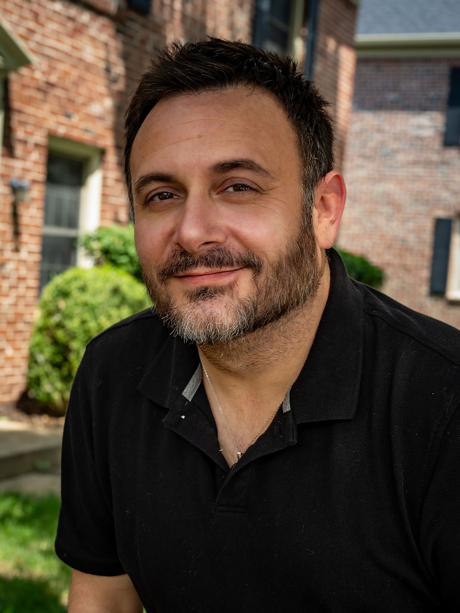 A smiling man with a beard wearing a black polo shirt stands outdoors near a brick building.
