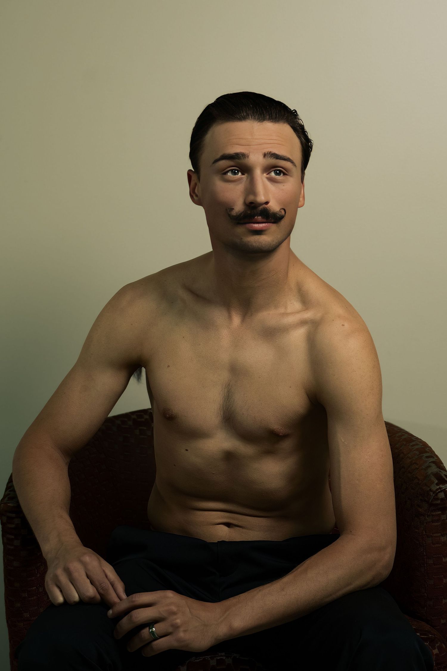 Shirtless muscular man with mustache sitting in armchair, looking up confidently against neutral background.