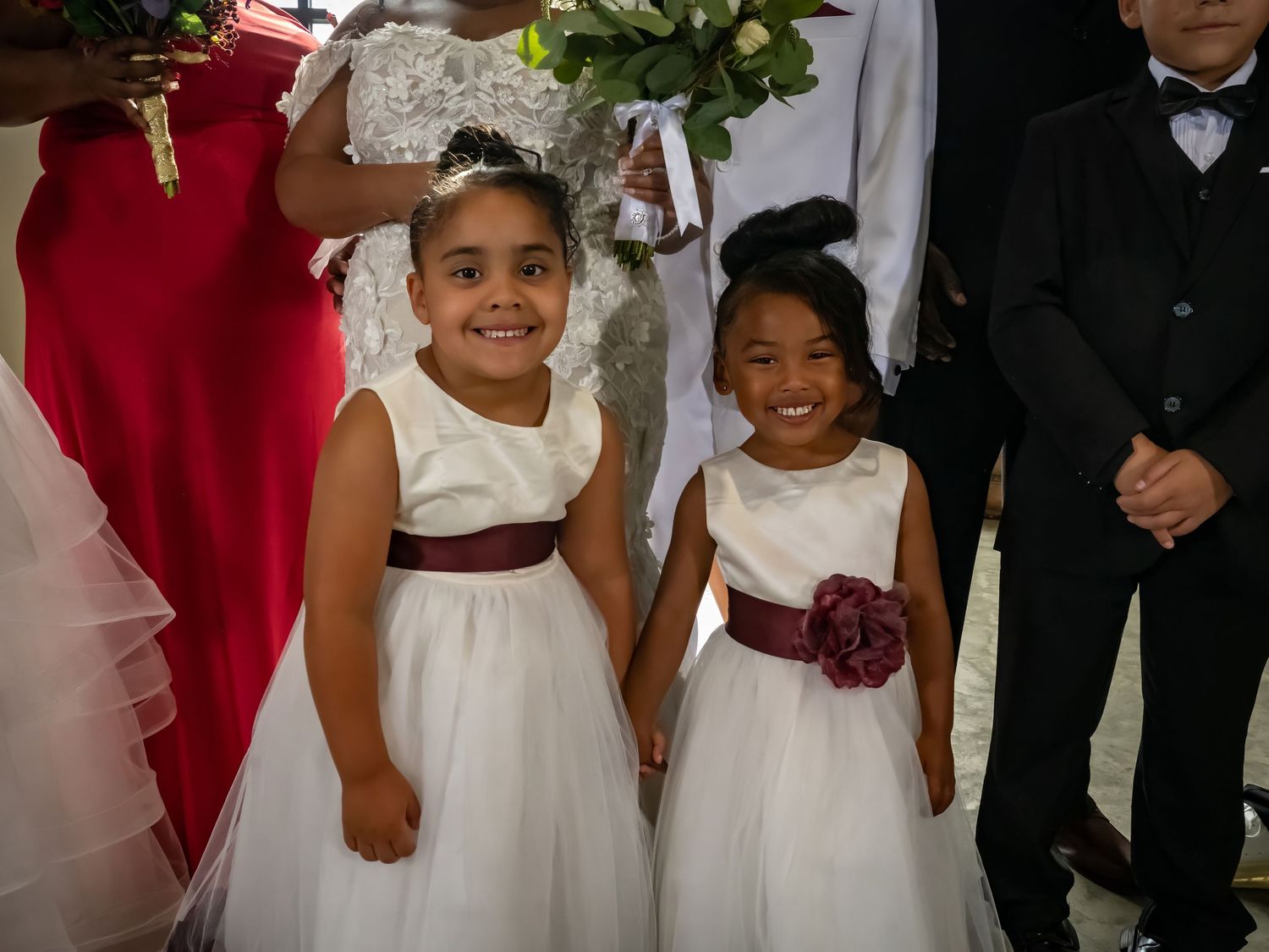 Children in formal white dresses and red accents stand together at a wedding.