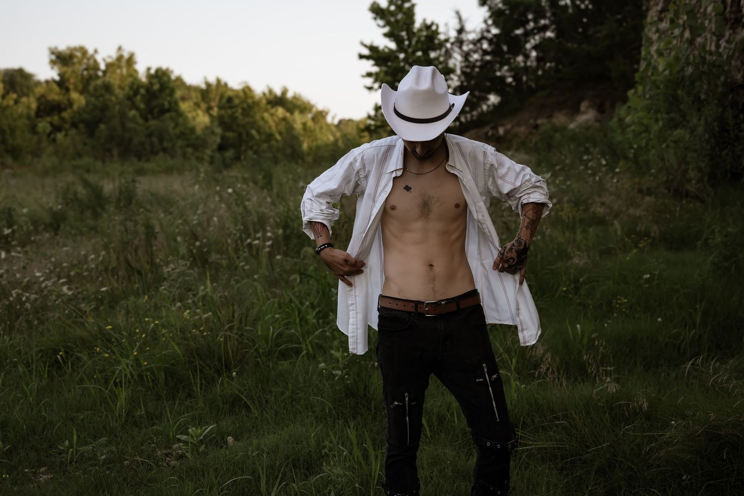 Outdoor portrait in white open shirt and cowboy hat at sunset in a grassy field.