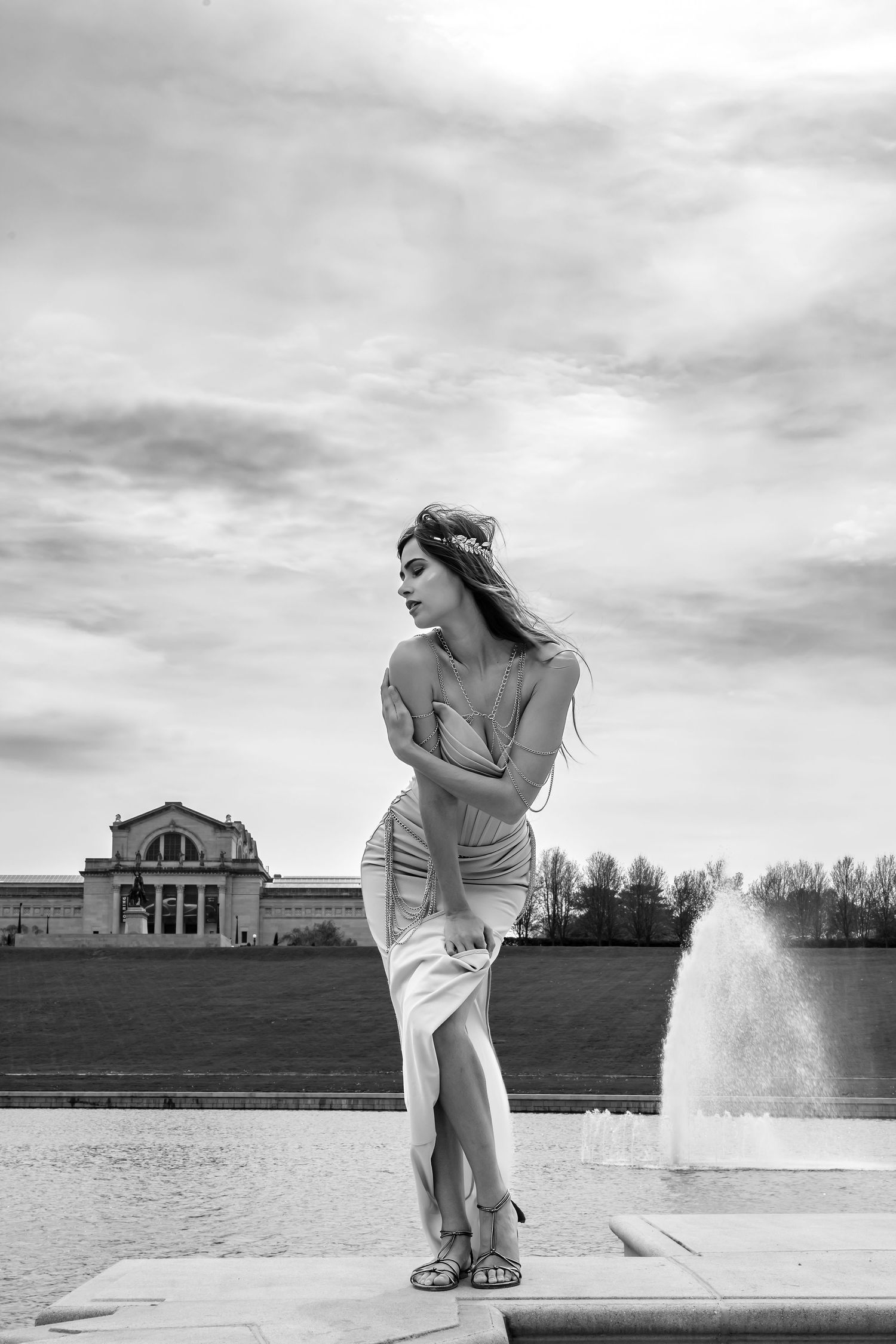 Black and white portrait of woman in casual dress posing by fountain with art museum in background.