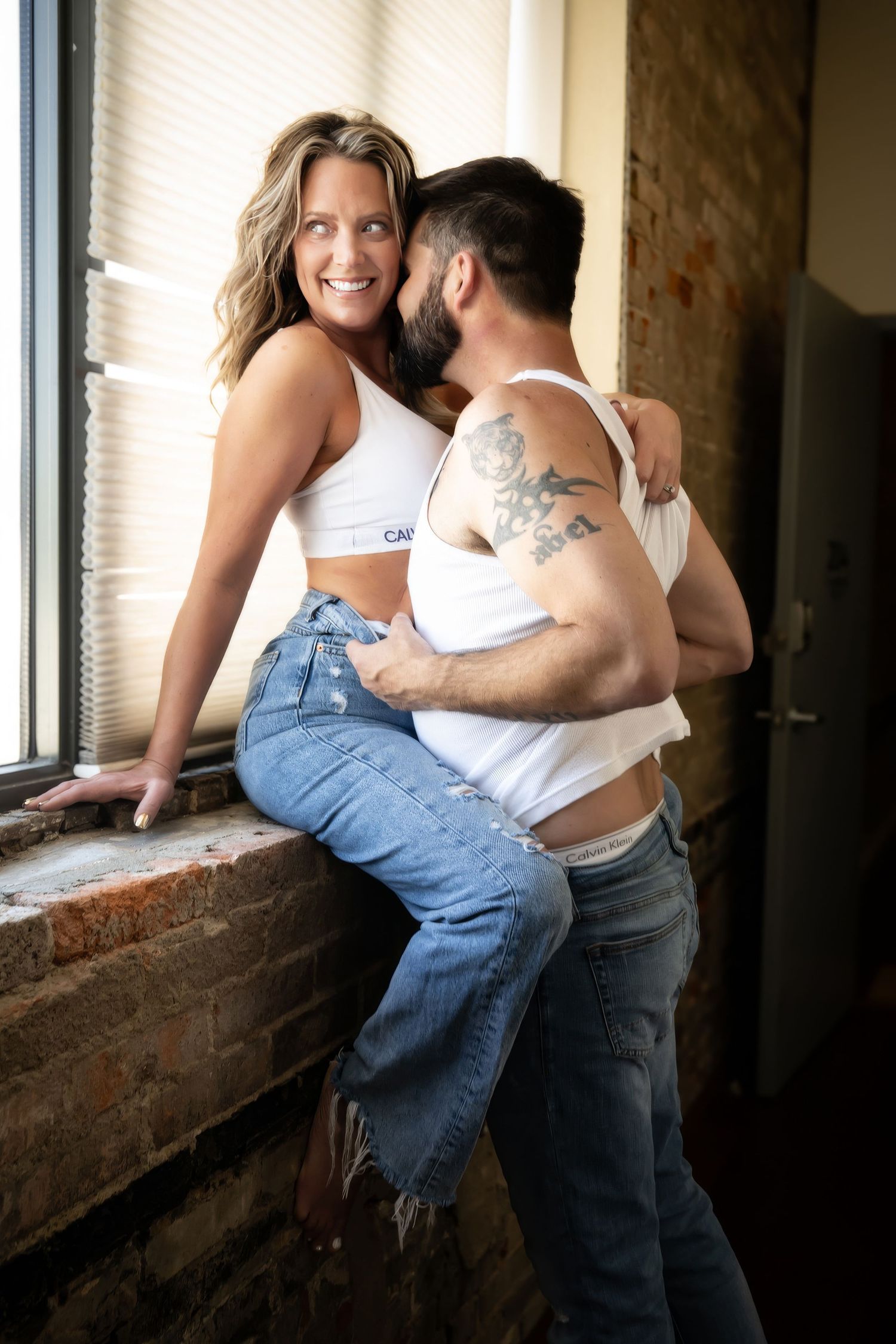 A couple embraces intimately near a window in a rustic industrial setting with exposed brick walls.