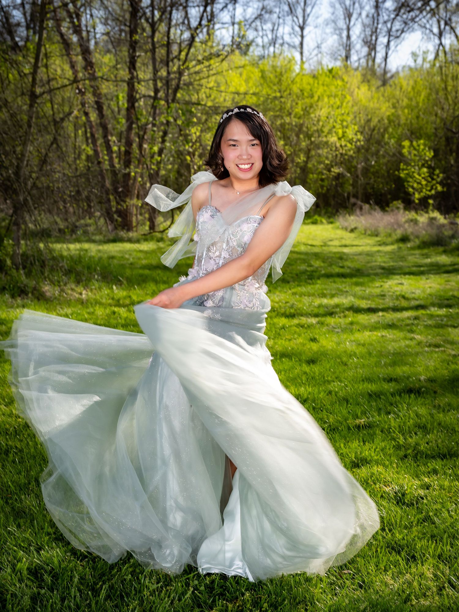 A flowing white wedding dress twirls gracefully in a sunny spring meadow surrounded by trees.