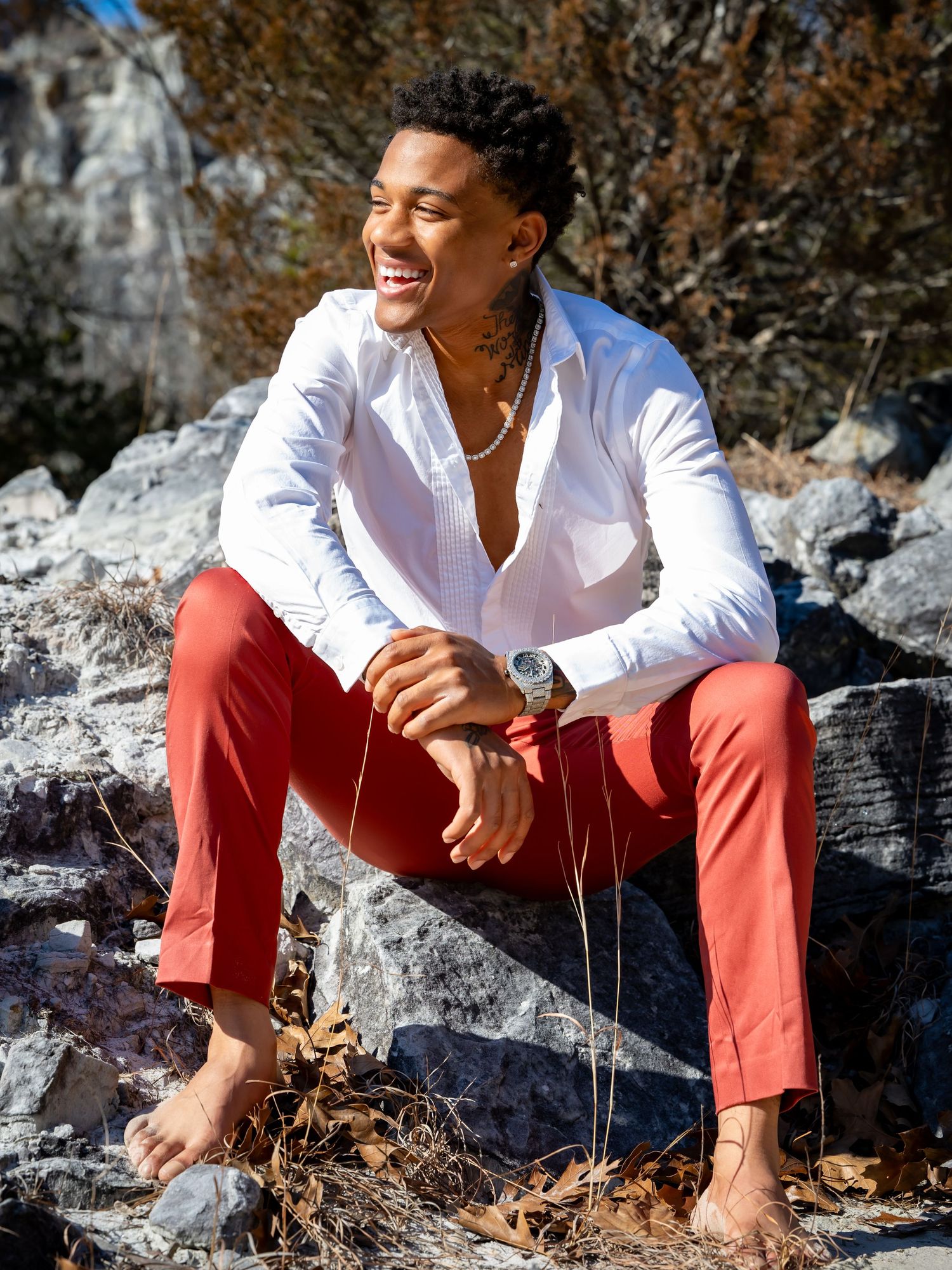 Young man smiling, wearing white shirt and red pants, sitting barefoot on rocks outdoors in sunny weather.