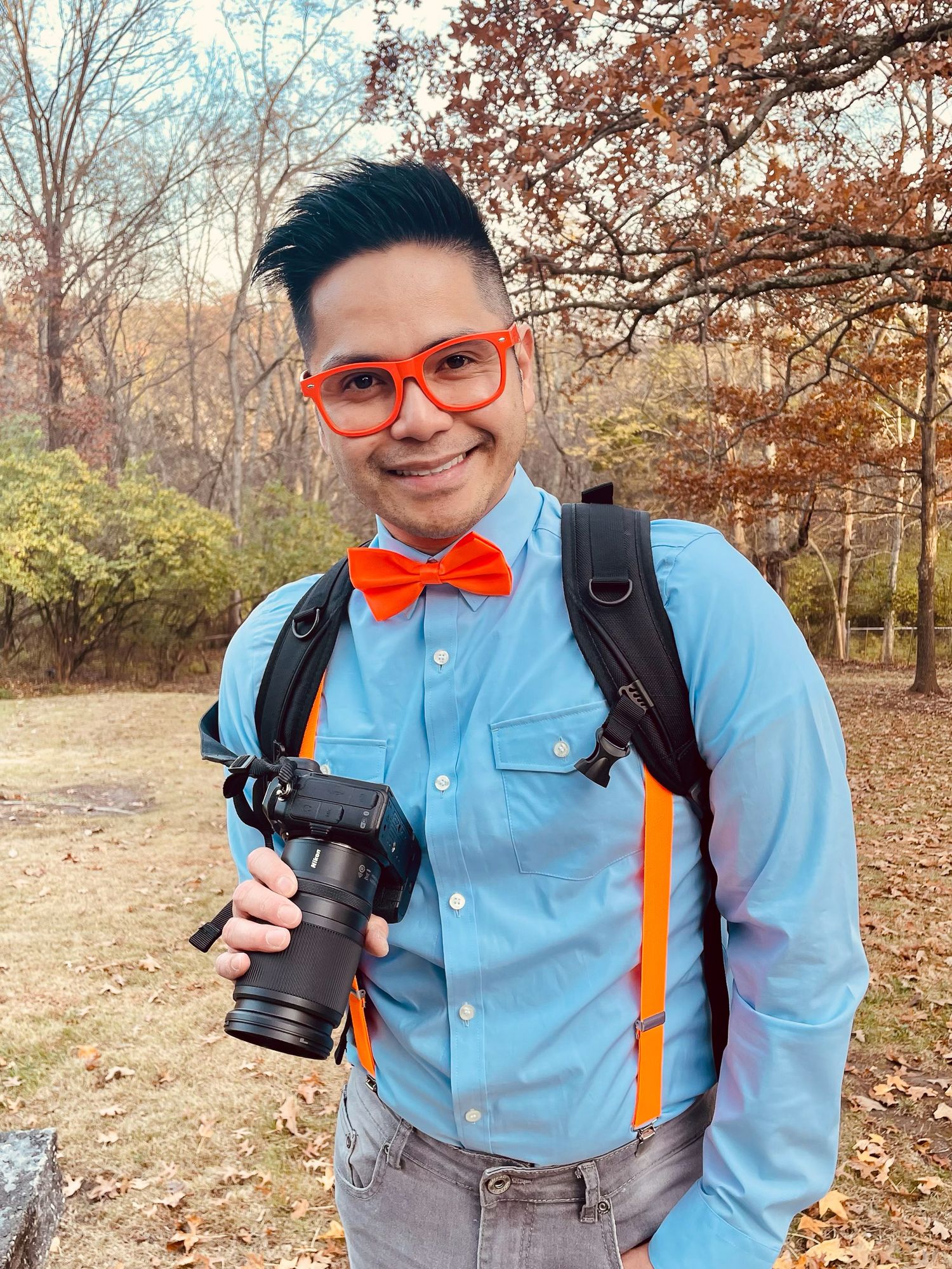 Man in blue shirt, orange bow tie, suspenders, and glasses holds a camera outdoors in autumn.