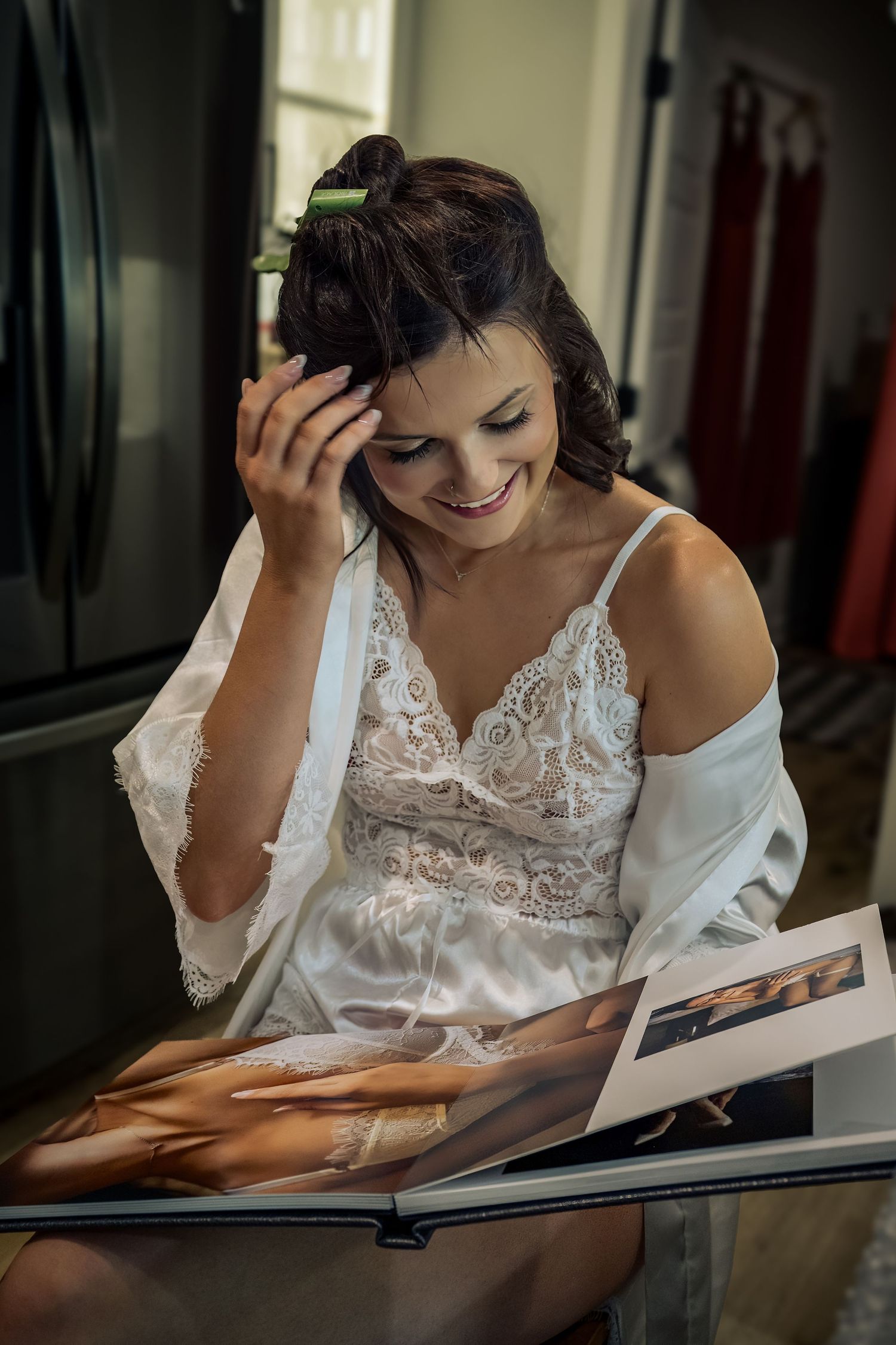 Smiling bride in white lace robe reads wedding album while fixing her hair before ceremony.