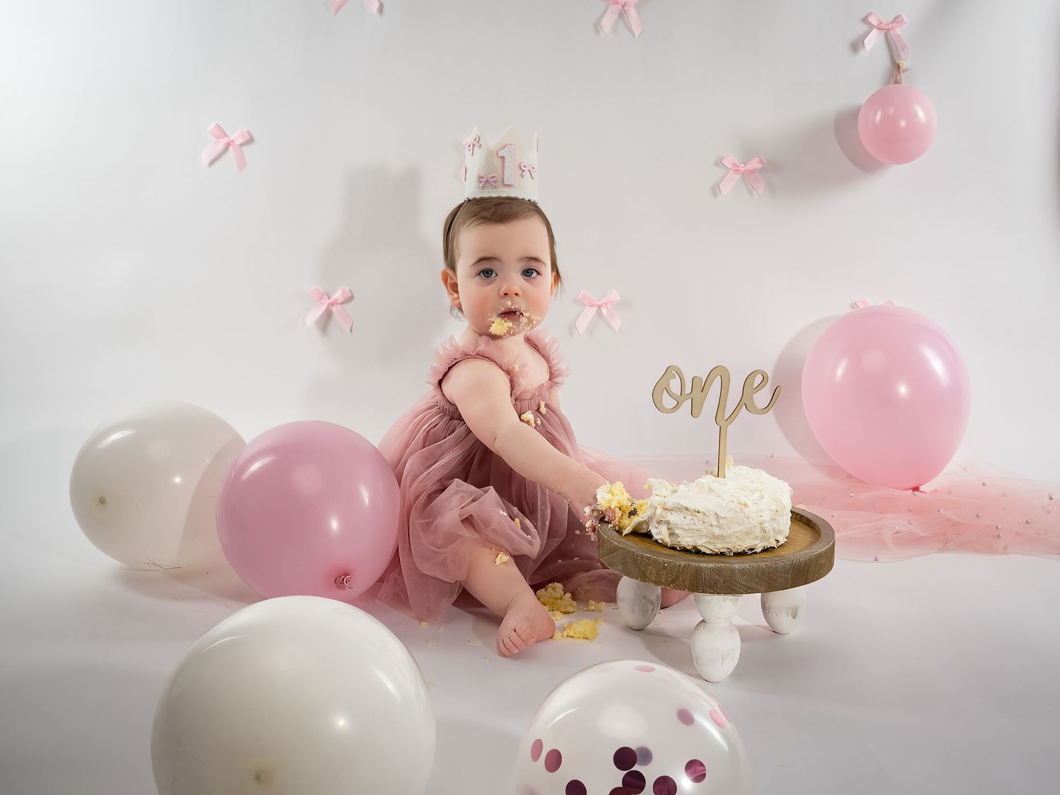 First birthday celebration setup with baby, cake, and pink and white balloons.